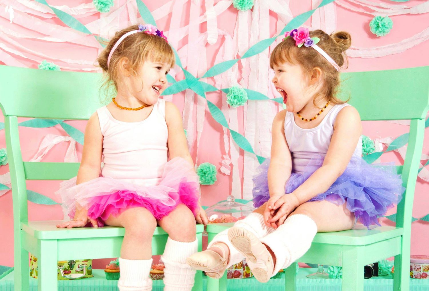 Two little girls in colorful tutus laughing together on mint green chairs at a festive pink party.