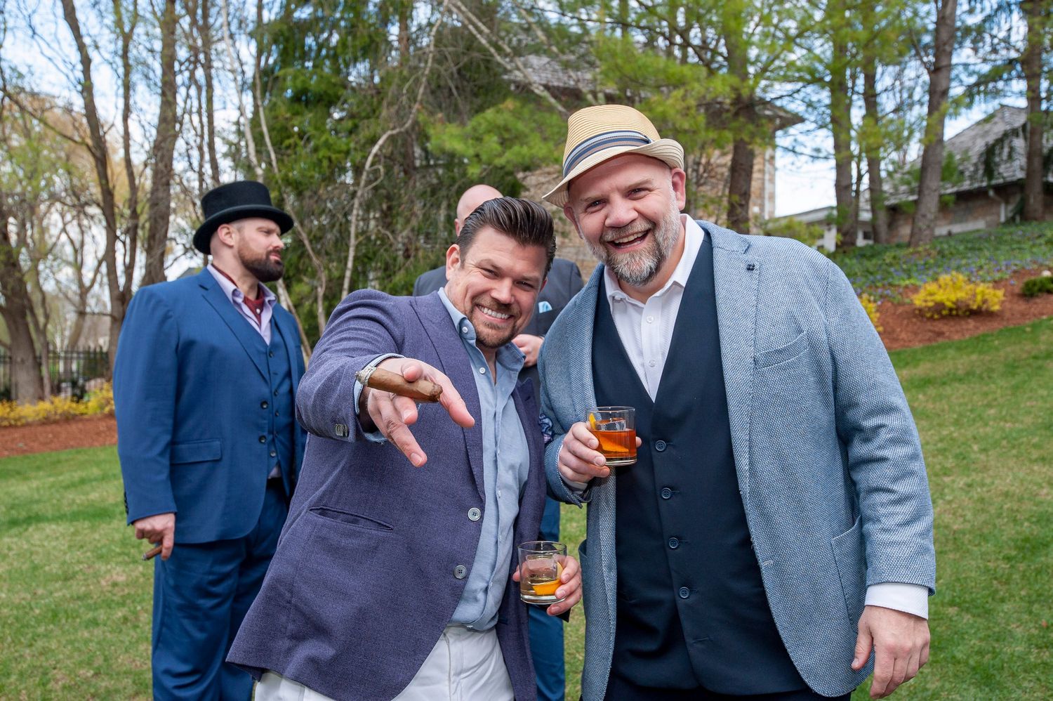 Three men in dapper suits laughing and enjoying drinks at an outdoor Kentucky Derby themed garden party.