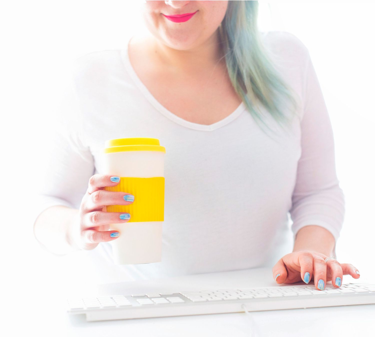 Woman with blue hair holding yellow coffee cup while typing on white keyboard.