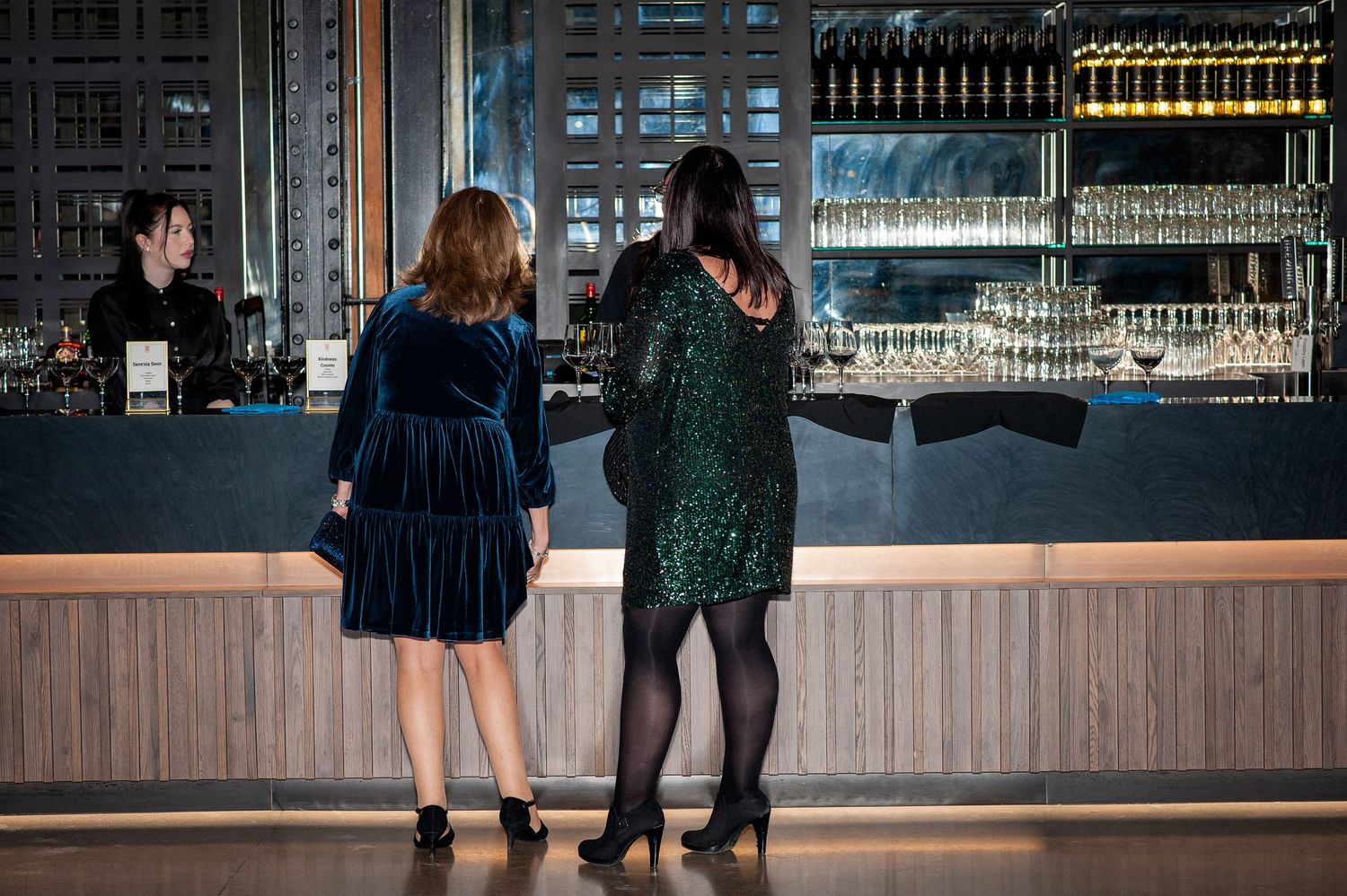 Two women in festive dresses standing at a modern bar with backlit glass shelves.