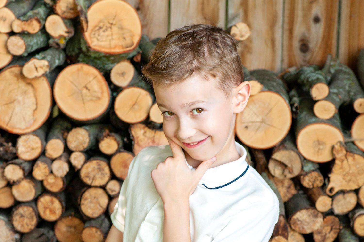Smiling young boy in white shirt posing confidently in front of stacked wooden logs.