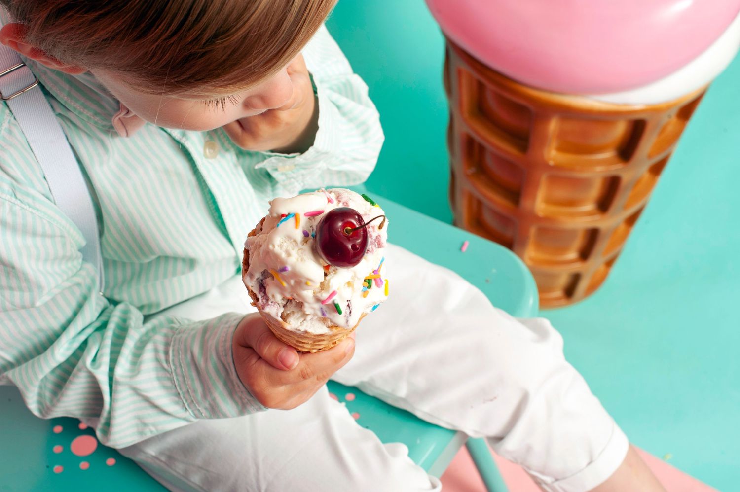 Young child holding a colorful ice cream cone with cherry on top, sitting near a large decorative ice cream prop.