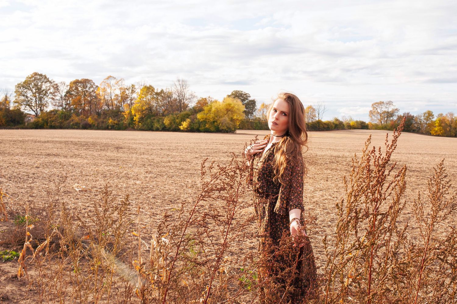 Woman in floral dress standing in a golden harvested field with autumn trees in background.