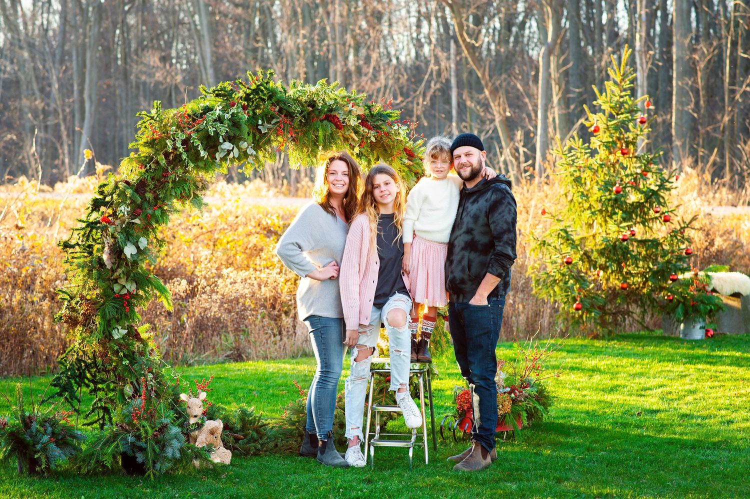 Family of four posing under a floral arch in an autumn outdoor setting with colorful fall foliage.