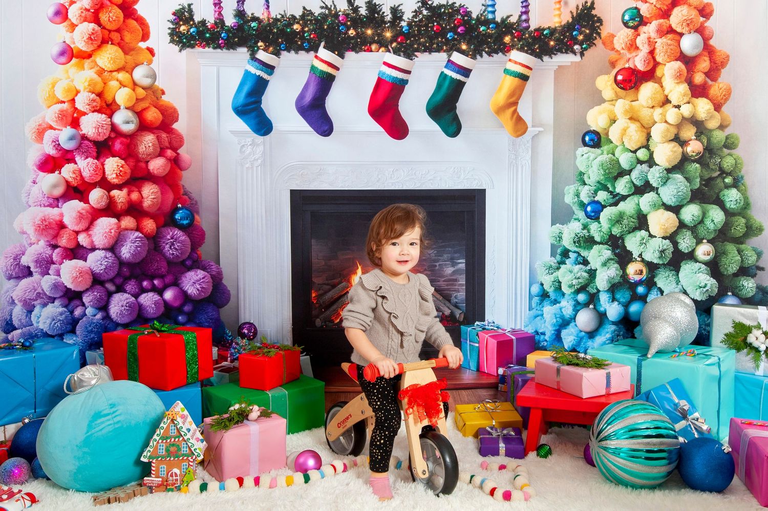 Toddler on balance bike surrounded by colorful gifts near rainbow Christmas trees and fireplace.