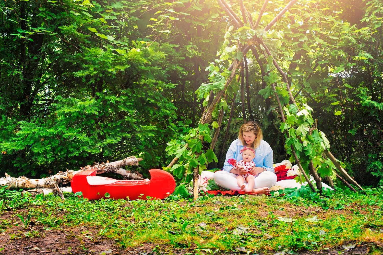Mother and baby sitting together outdoors near a teepee structure surrounded by lush green trees.
