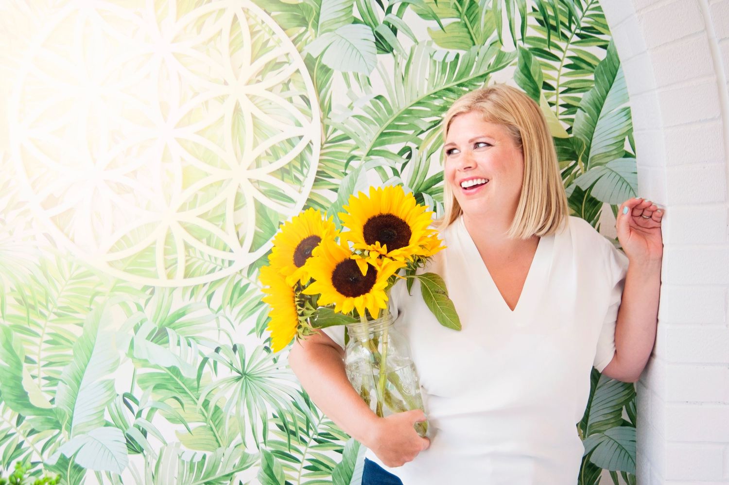 Joyful blonde woman holding sunflowers in vase against tropical leaf mural background.
