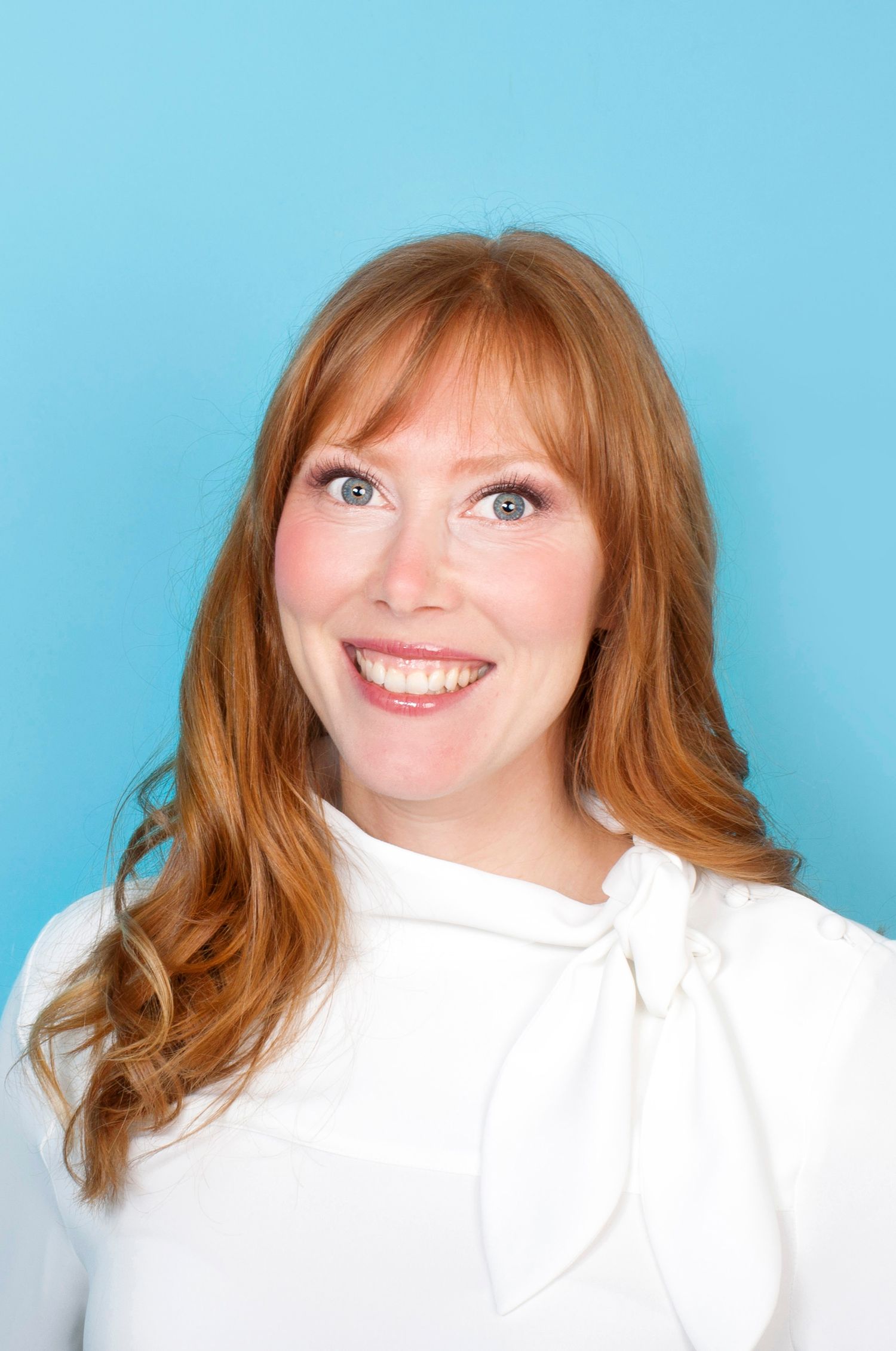 Smiling redhead woman in white bow-neck blouse against bright blue background.