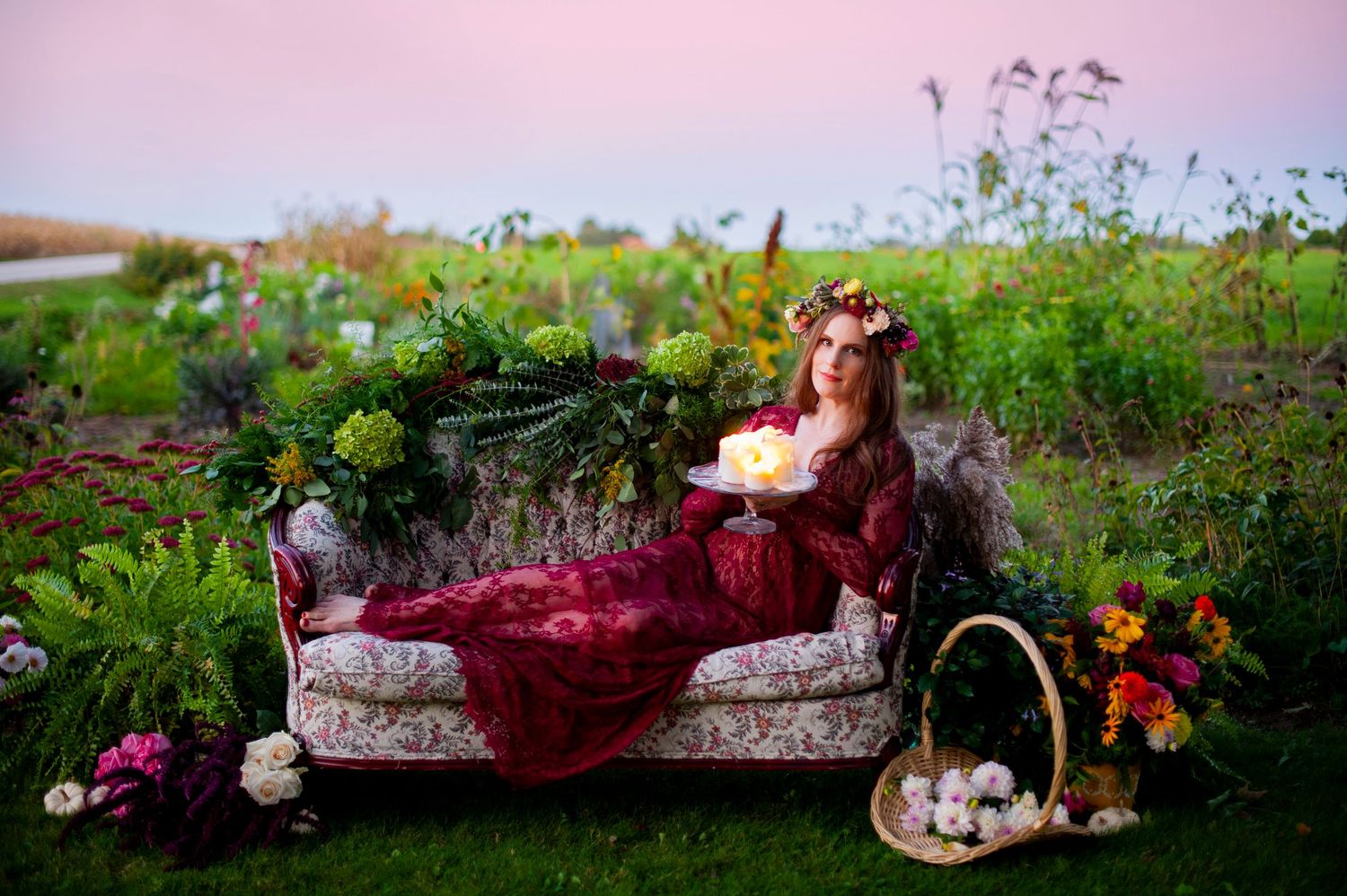 Woman in burgundy gown reclining on vintage sofa surrounded by flowers and greenery at sunset.