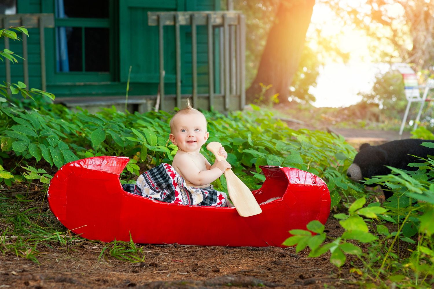 Happy baby sitting in a red toy canoe with a paddle outdoors surrounded by green foliage.