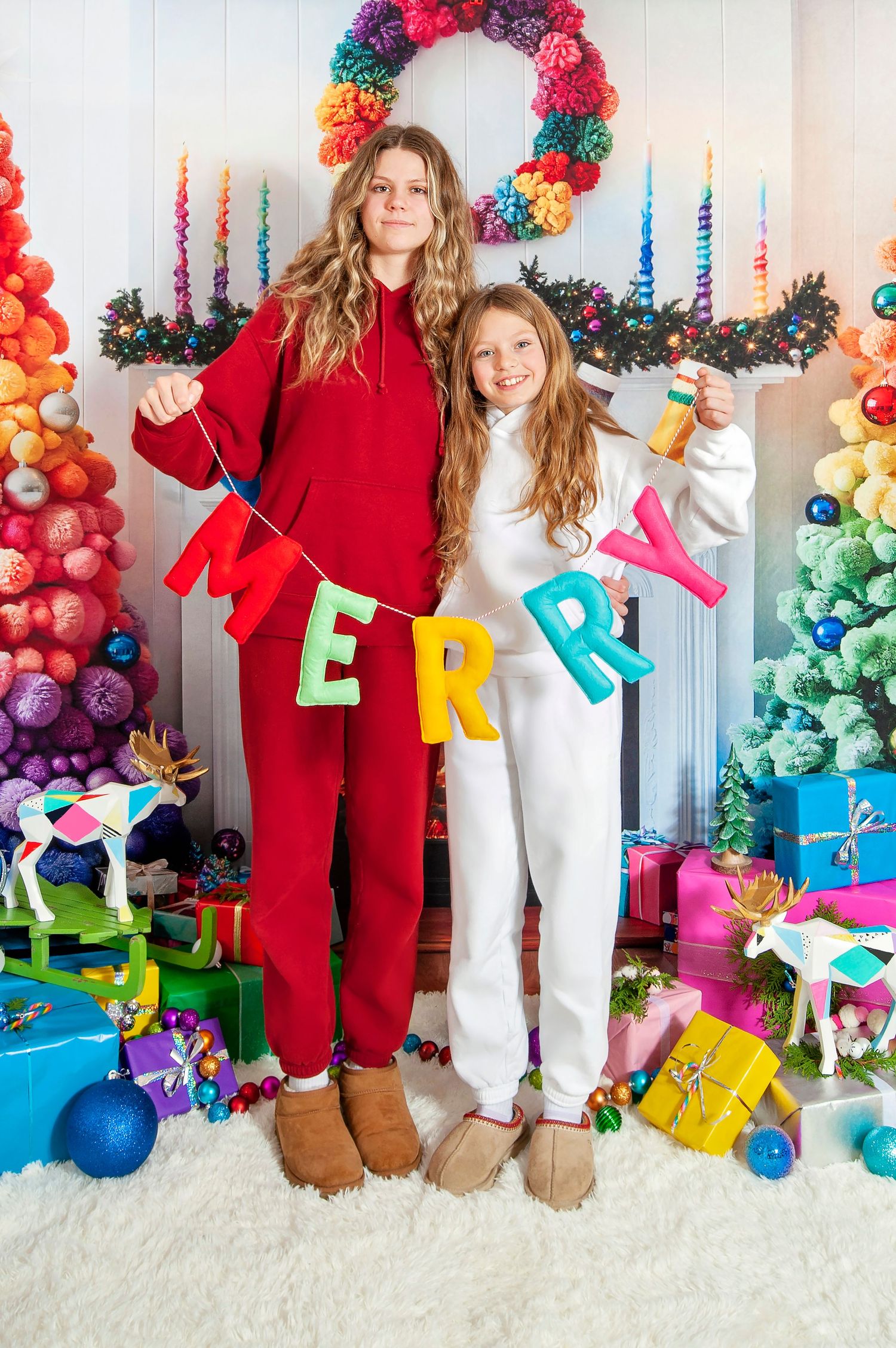 Two girls holding colorful MERRY banner in front of rainbow Christmas tree with gifts.