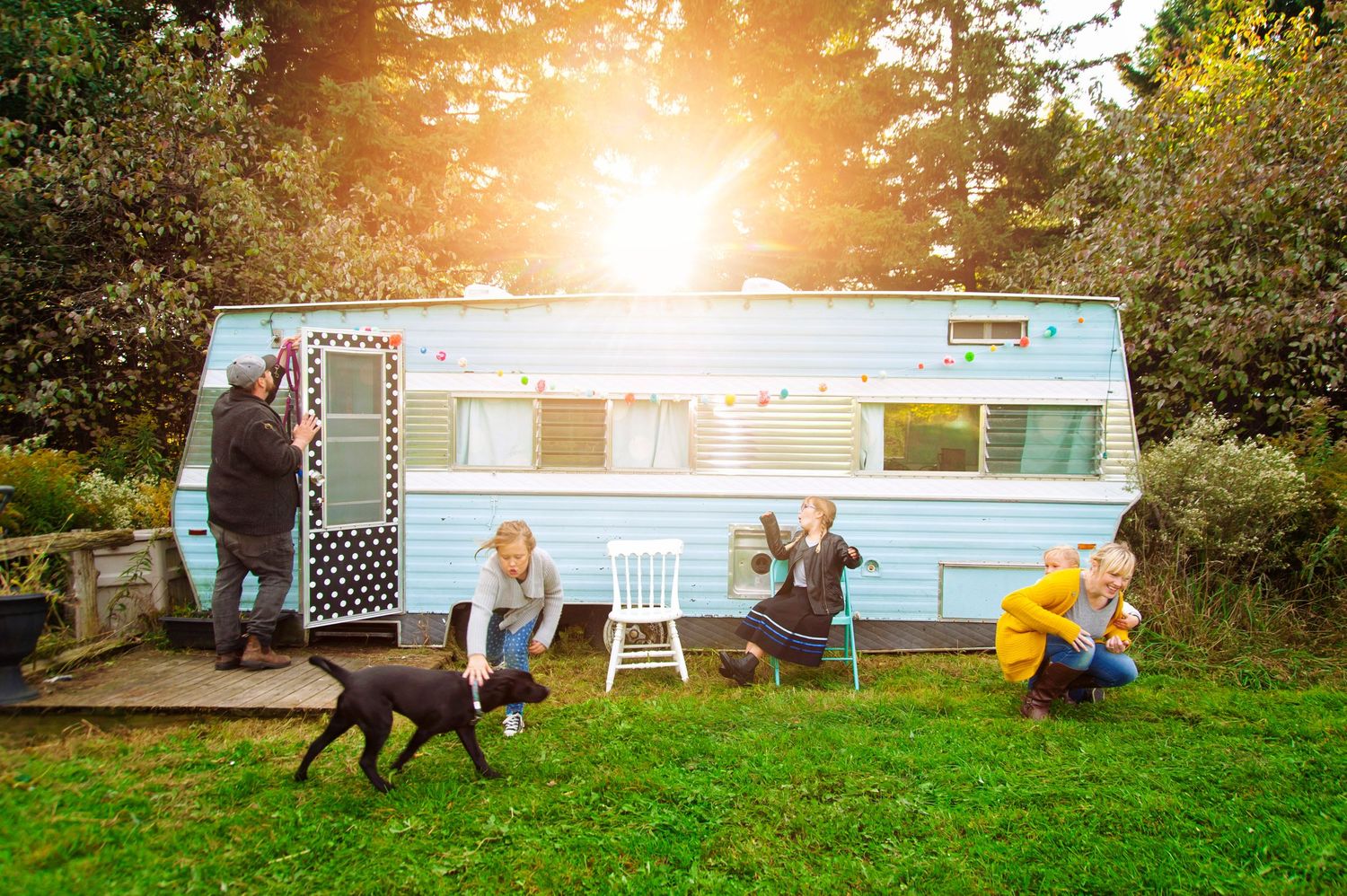 Family playing outside vintage blue caravan with black dog on green lawn in golden sunlight.