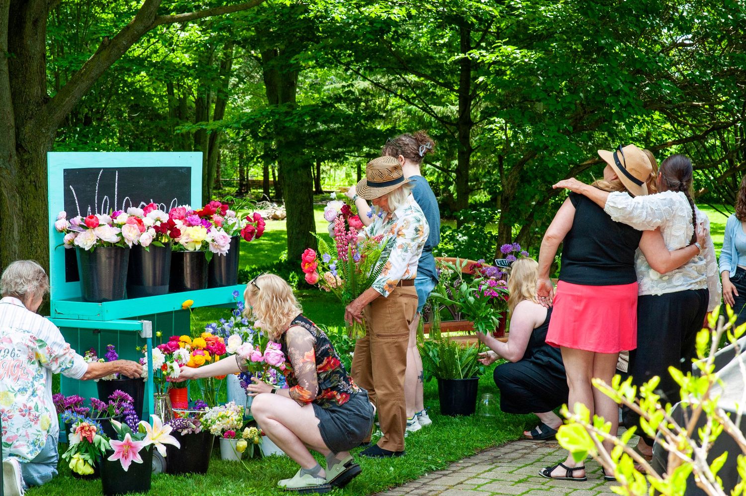 People gathering and hugging outdoors at a colorful flower market event surrounded by lush green trees.