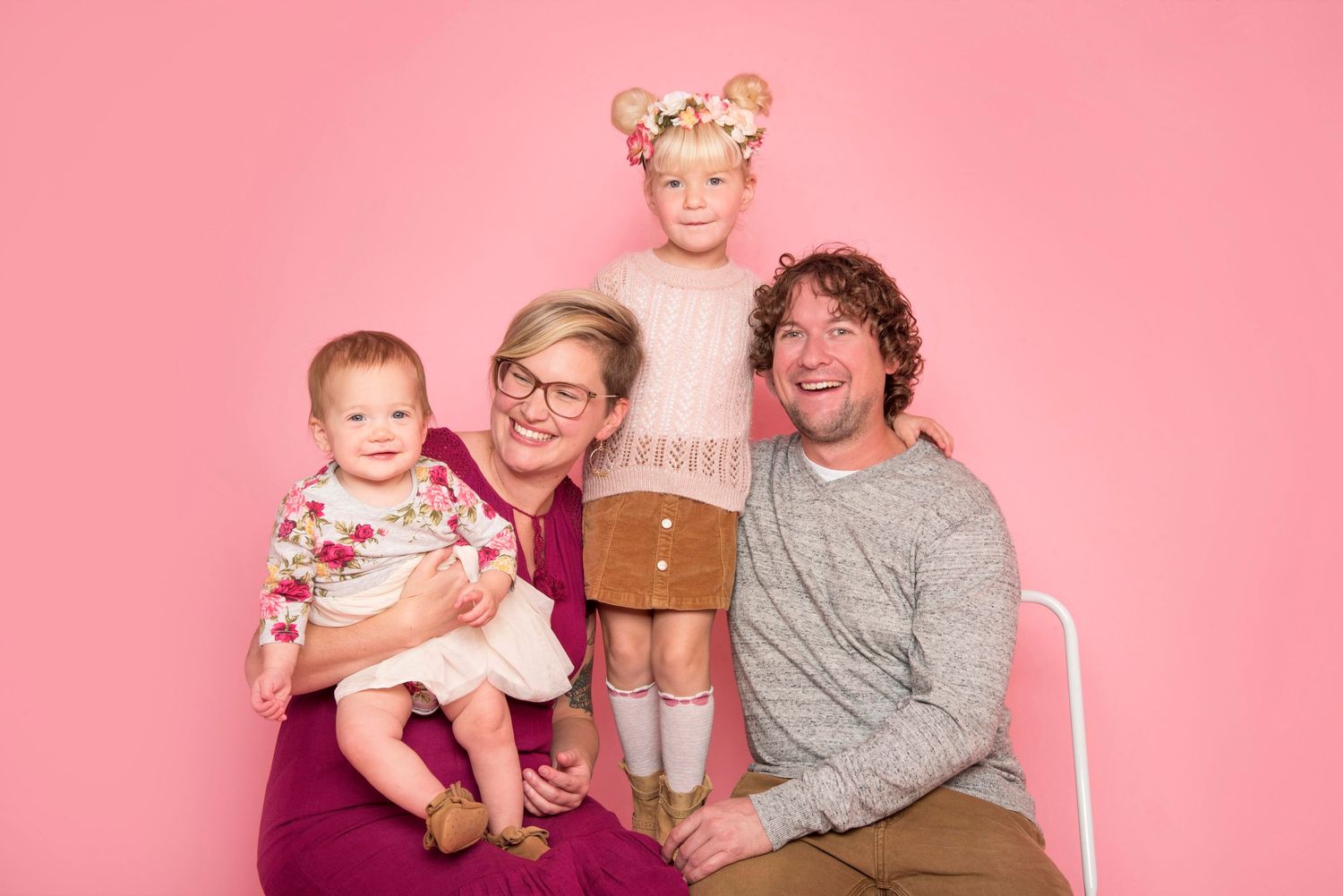 Happy family of four with two young daughters posing together against a cheerful pink studio background.