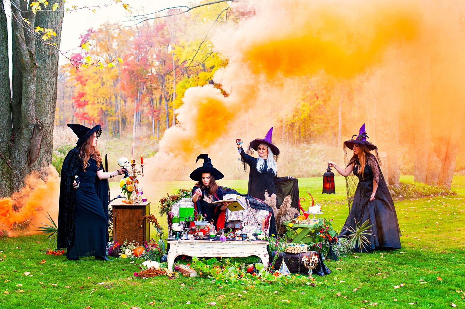 Five women in witch costumes gather around a Halloween table outdoors with orange smoke bombs in autumn setting.