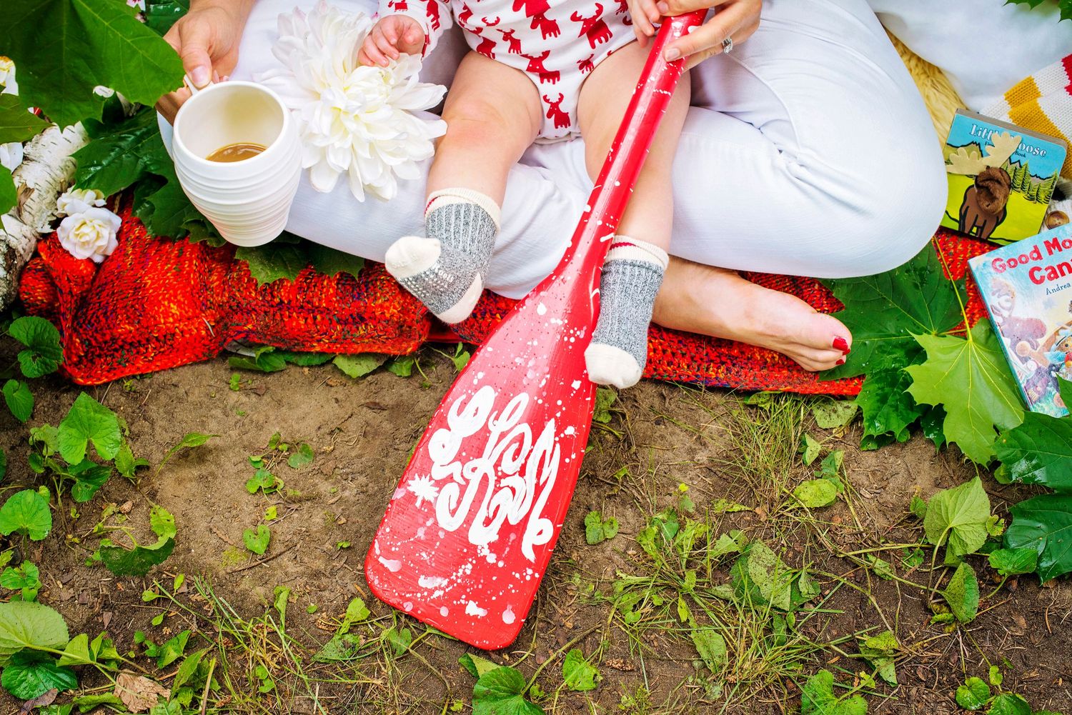 Mother and baby sitting outdoors on red blanket with a red paddle, cup, and colorful books nearby.