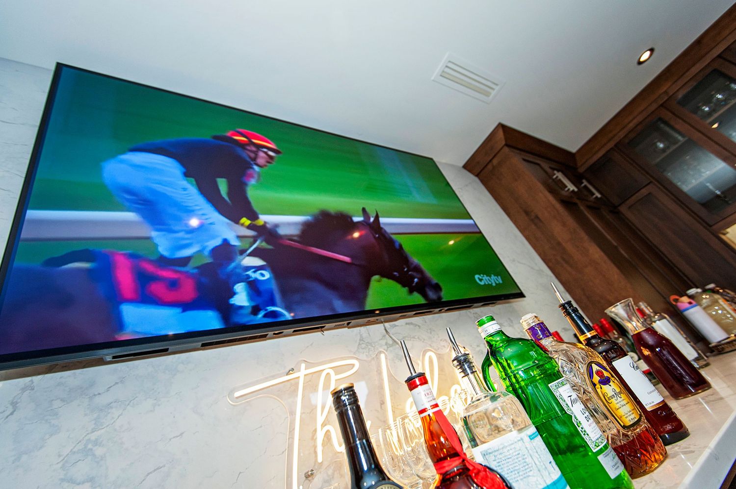 Large TV screen showing horse racing above a fully stocked bar at a Kentucky Derby watch party.