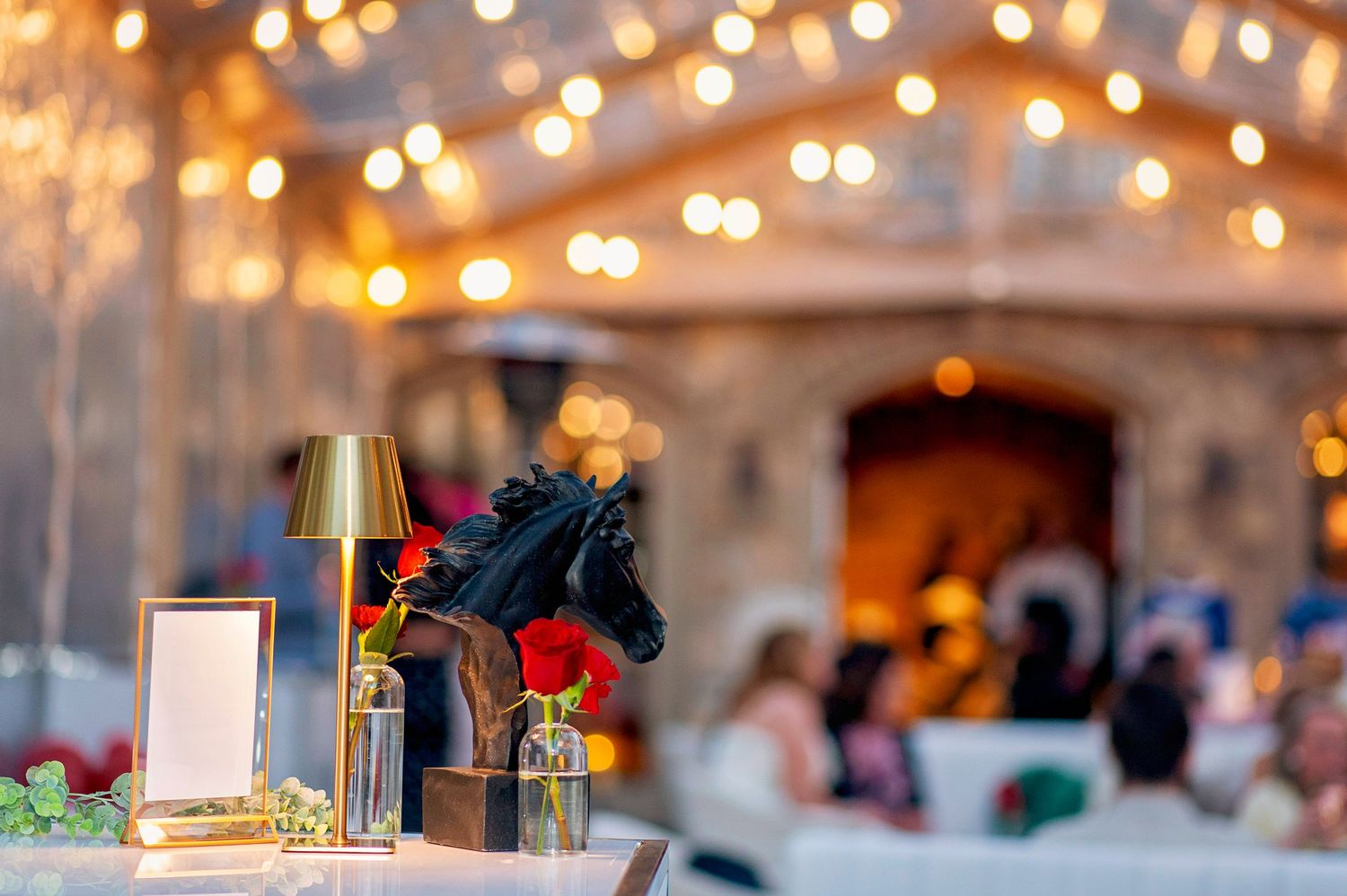 Elegant event decor with gold lamp, black horse figurine, and bokeh string lights at an outdoor venue.
