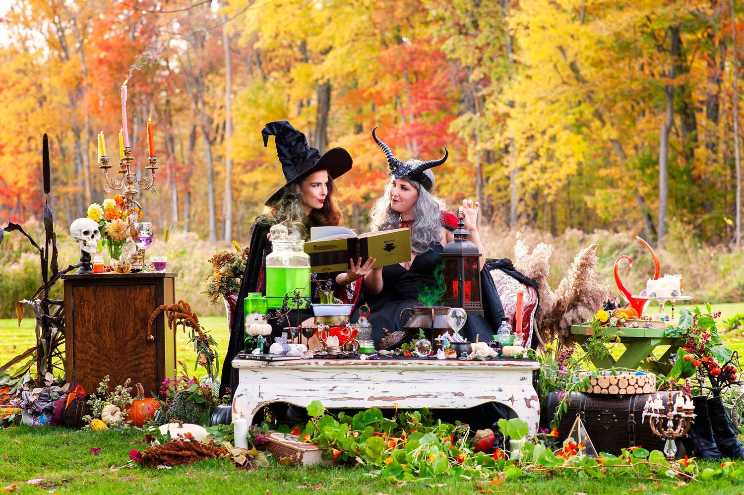 Two women in witch costumes reading a spell book outdoors amid Halloween decorations with colorful autumn foliage.