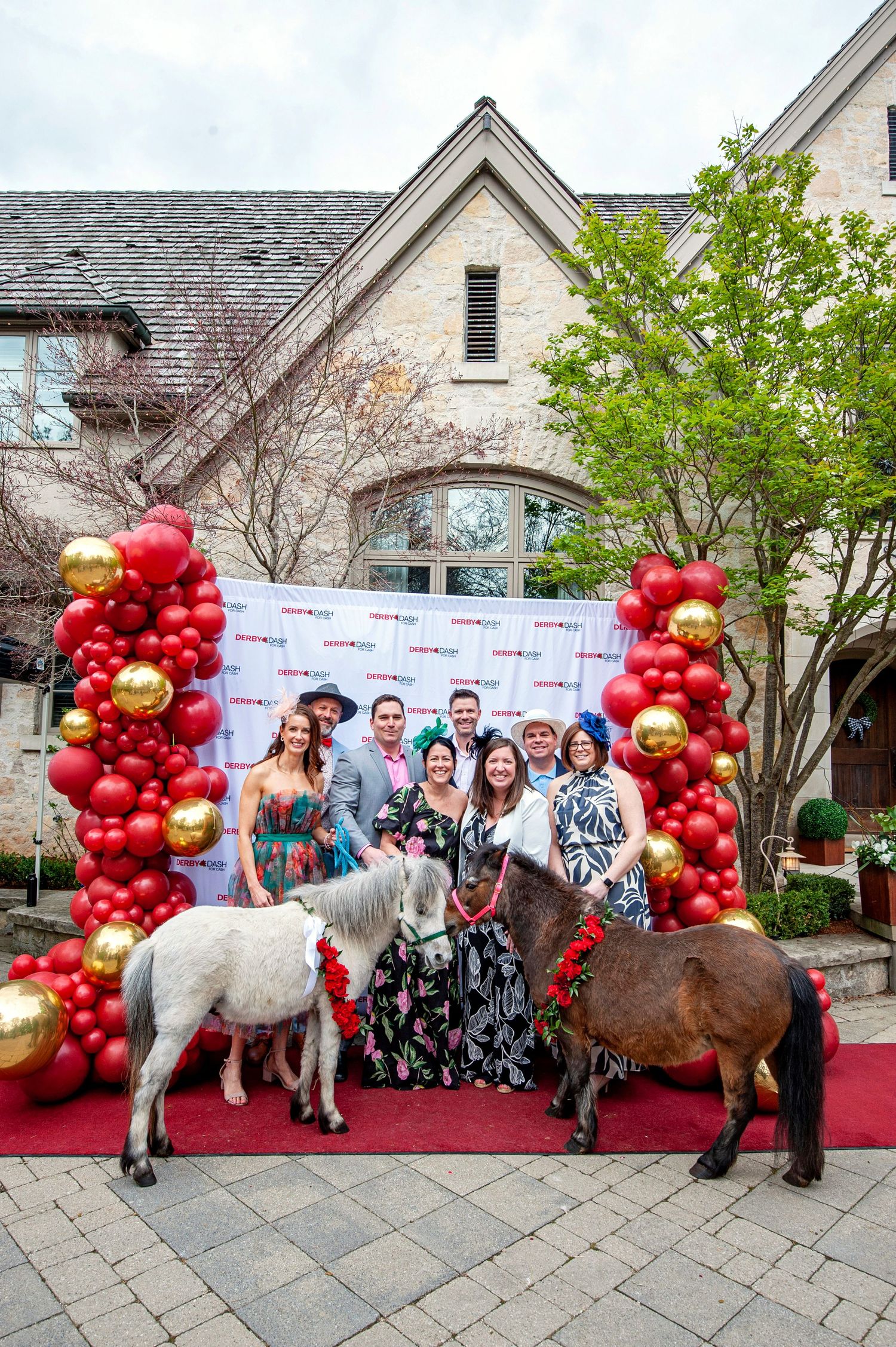 Group of elegantly dressed guests posing with miniature horses at a Kentucky Derby themed party with red balloons.