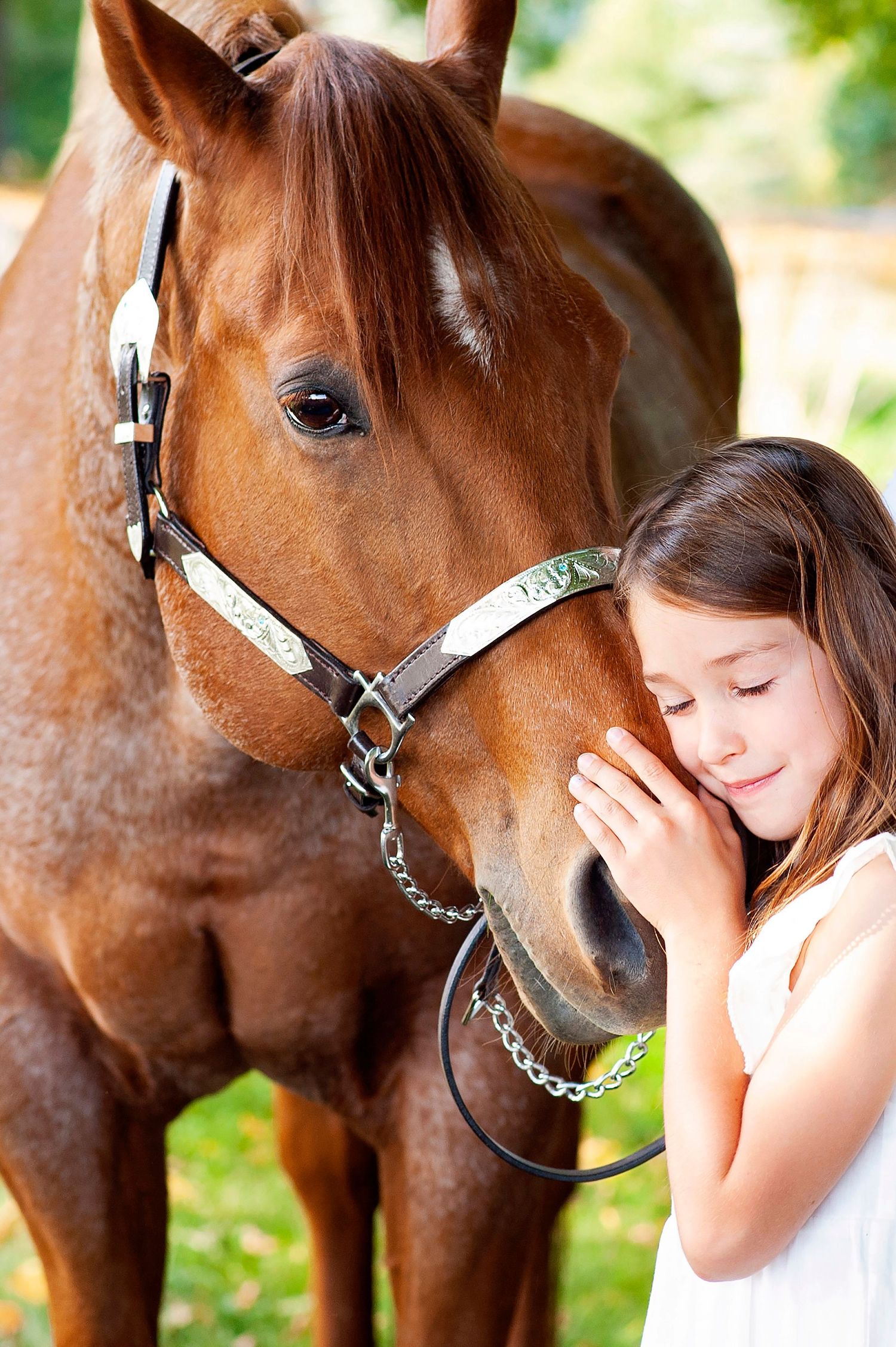 Young girl with closed eyes tenderly holding face of brown horse with silver bridle outdoors.