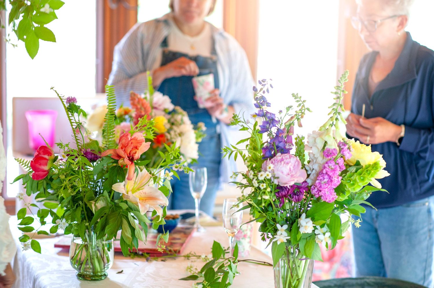 Colorful floral arrangements in glass vases on a table with women in the background at a flower workshop.