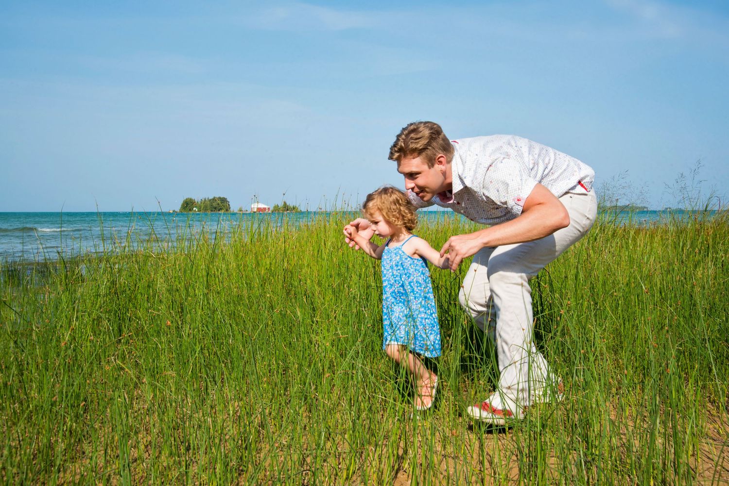 Father bending down to interact with toddler child in tall green grass near a scenic blue lake on a sunny day.