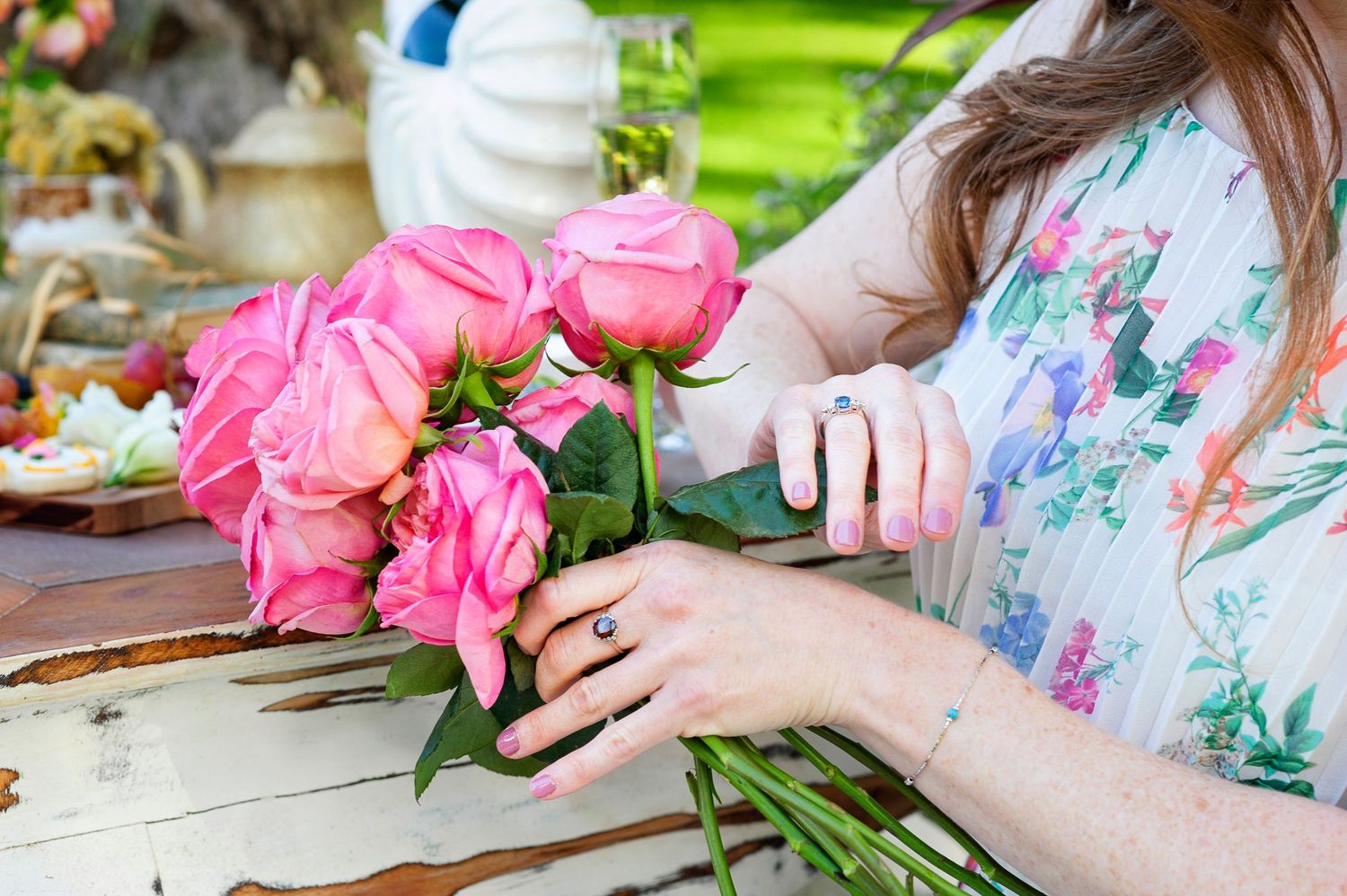 Woman in floral dress arranging pink roses bouquet on rustic wooden table outdoors.