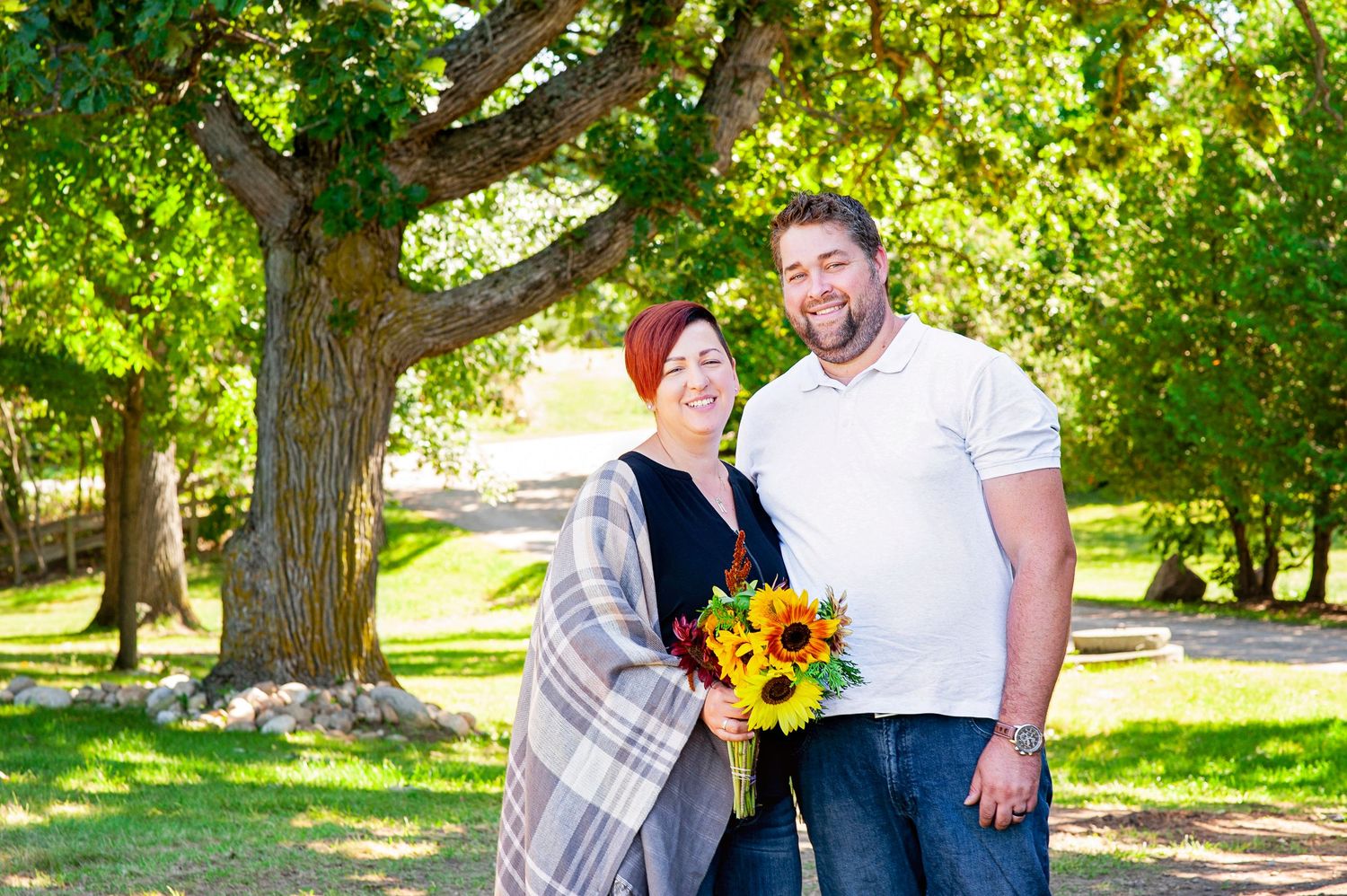 Couple smiling outdoors under large tree holding sunflower bouquet in park.