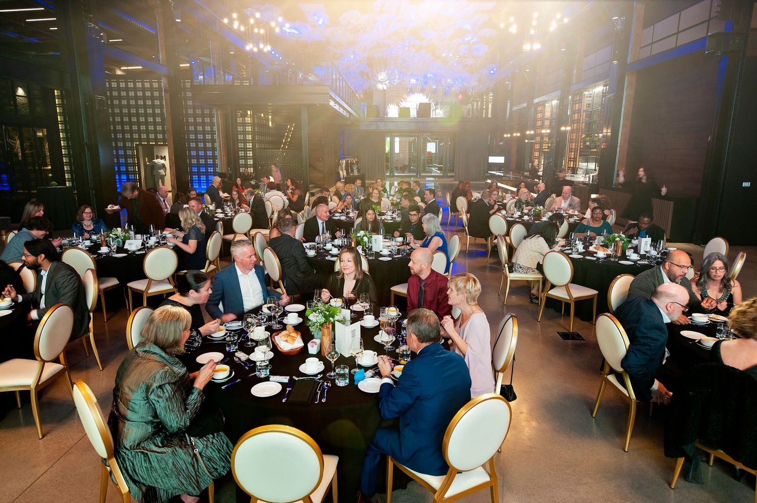 Elegant gala dinner event with guests seated at round black-draped tables in a grand venue.