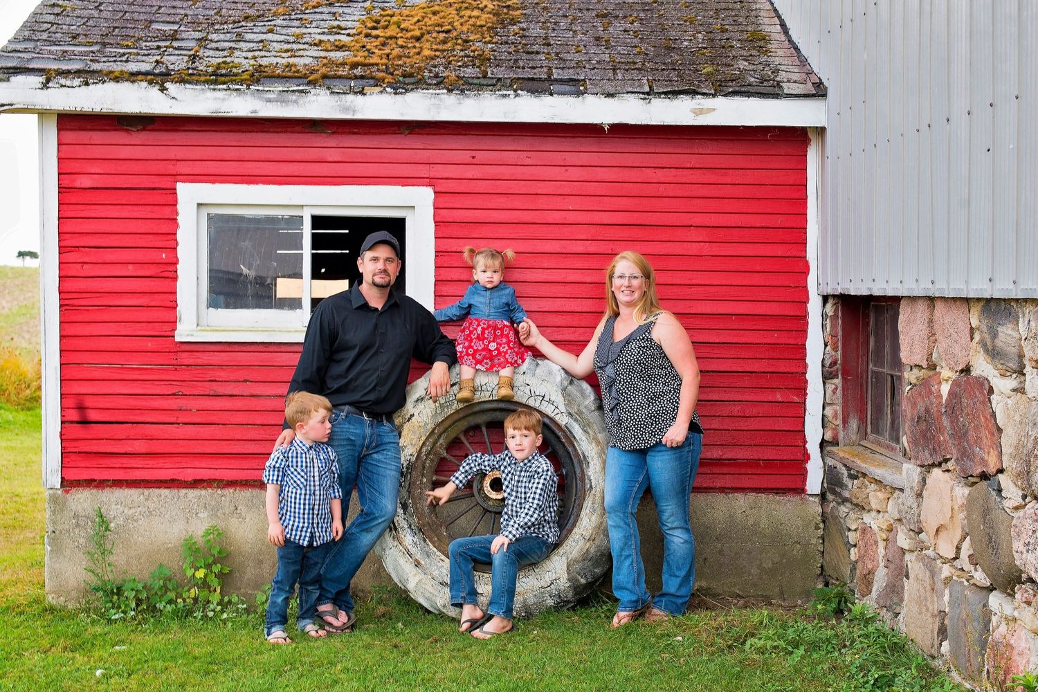 Happy family of five posing by a red barn with a large tire on a farm.