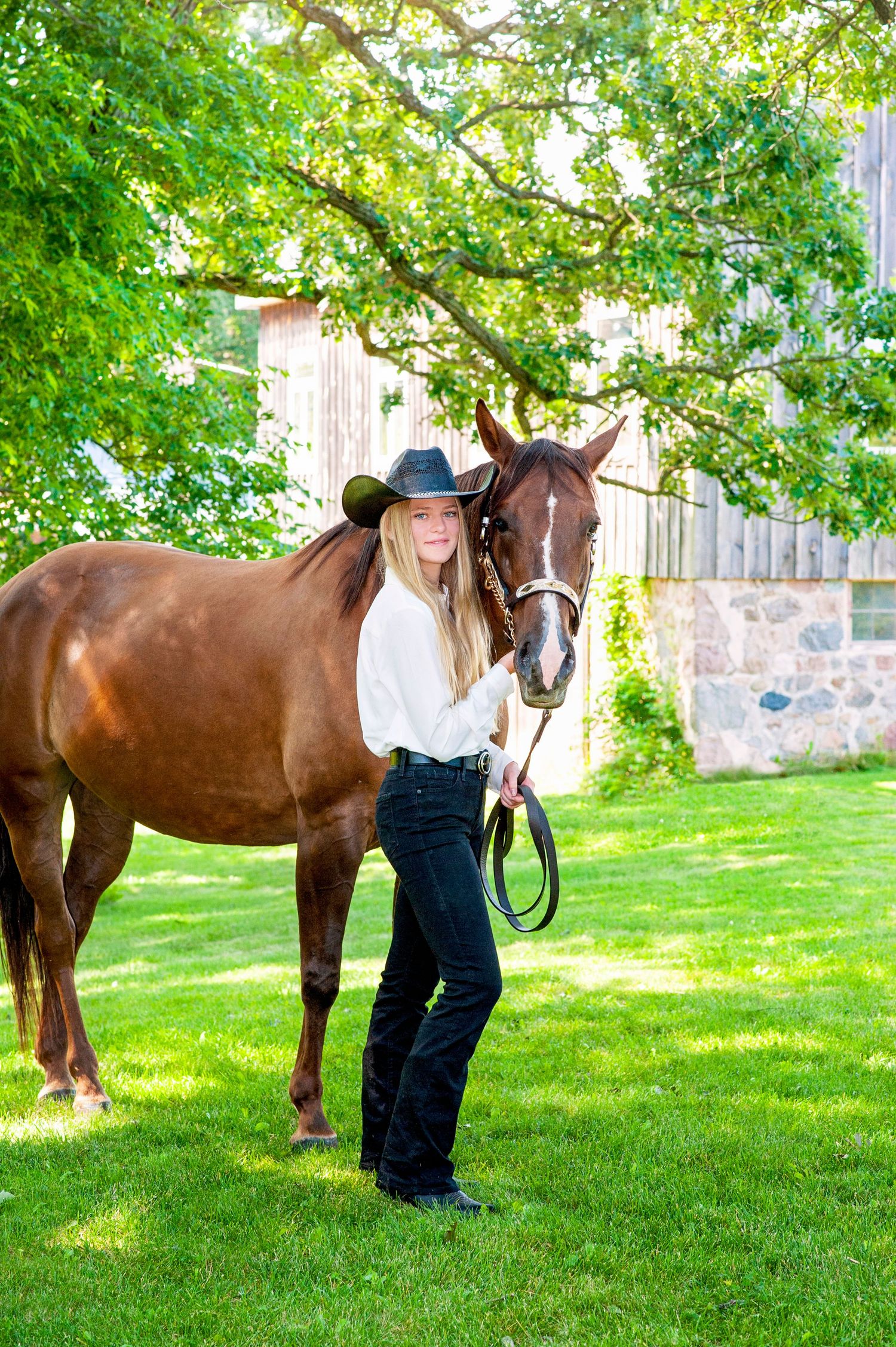 Young blonde woman in cowboy hat standing beside chestnut horse on green lawn.
