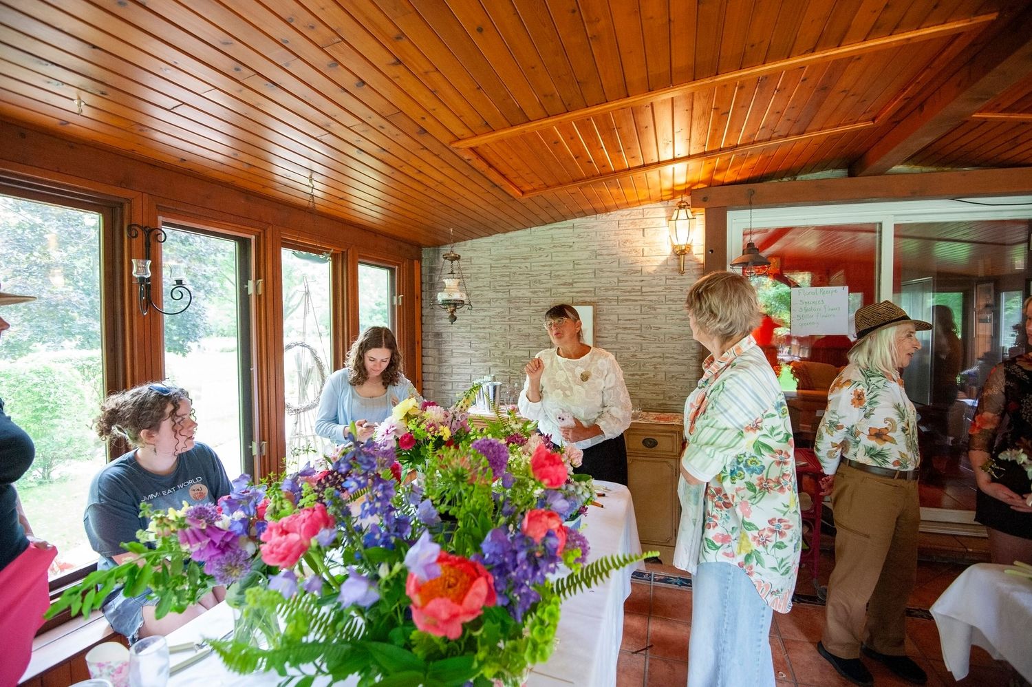 Guests mingling in a warm wooden sunroom with a large colorful floral centerpiece on the table.