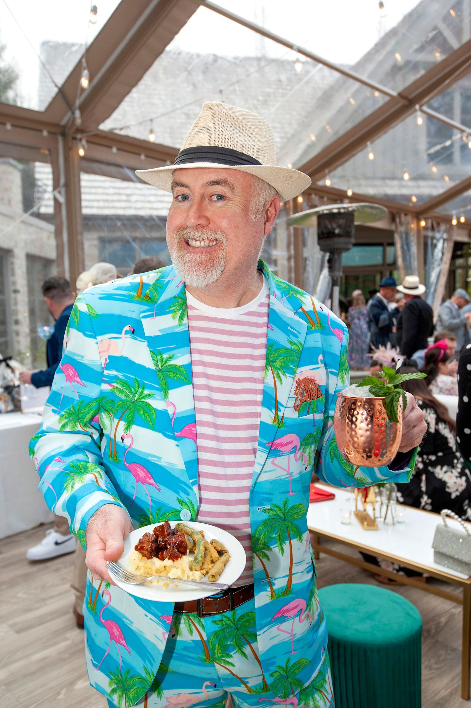 Man in tropical flamingo suit holding food and copper Moscow Mule mug at an outdoor Kentucky Derby event.
