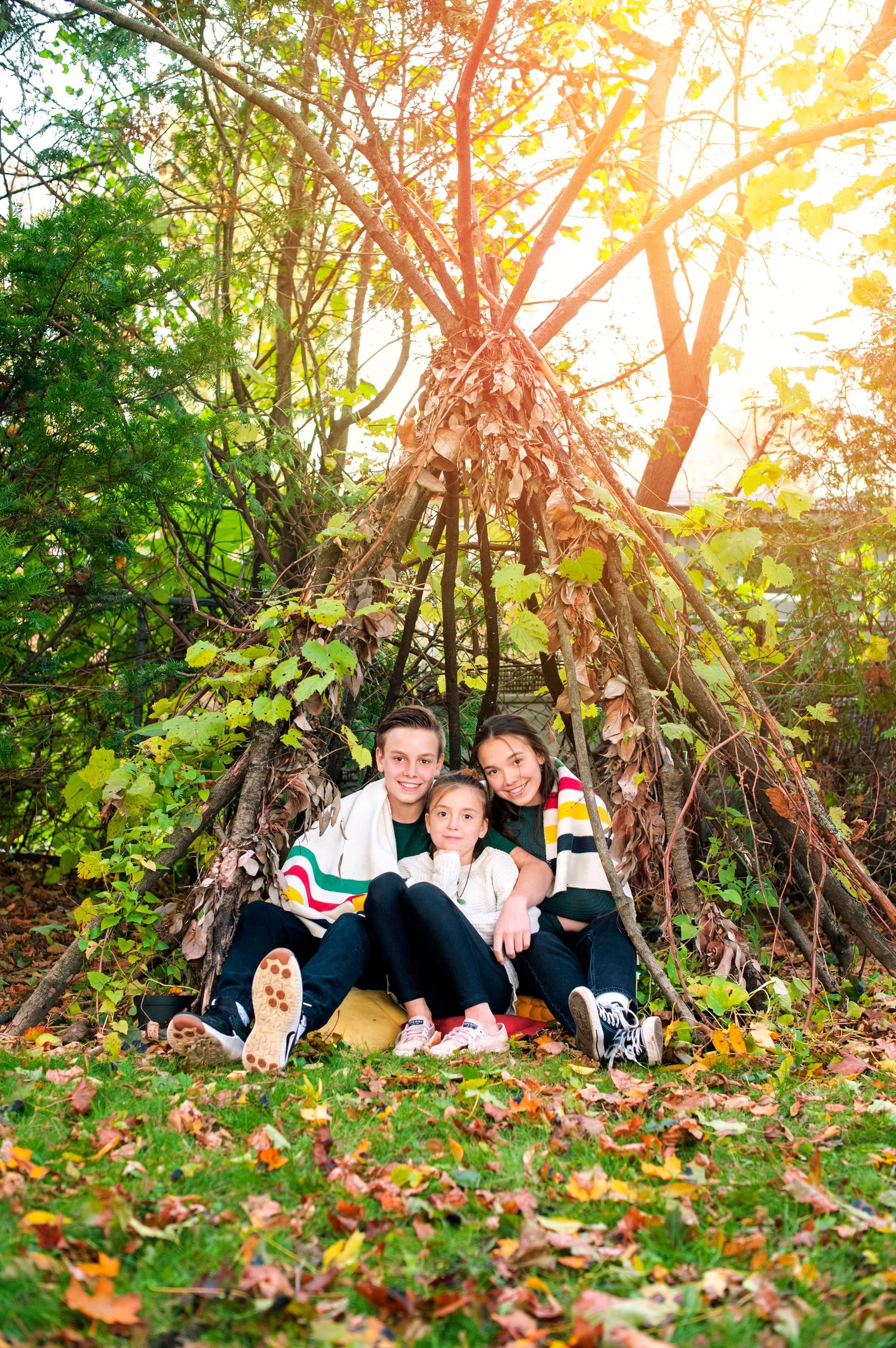 Family of three sitting together in front of a rustic teepee made of branches in an autumn forest.