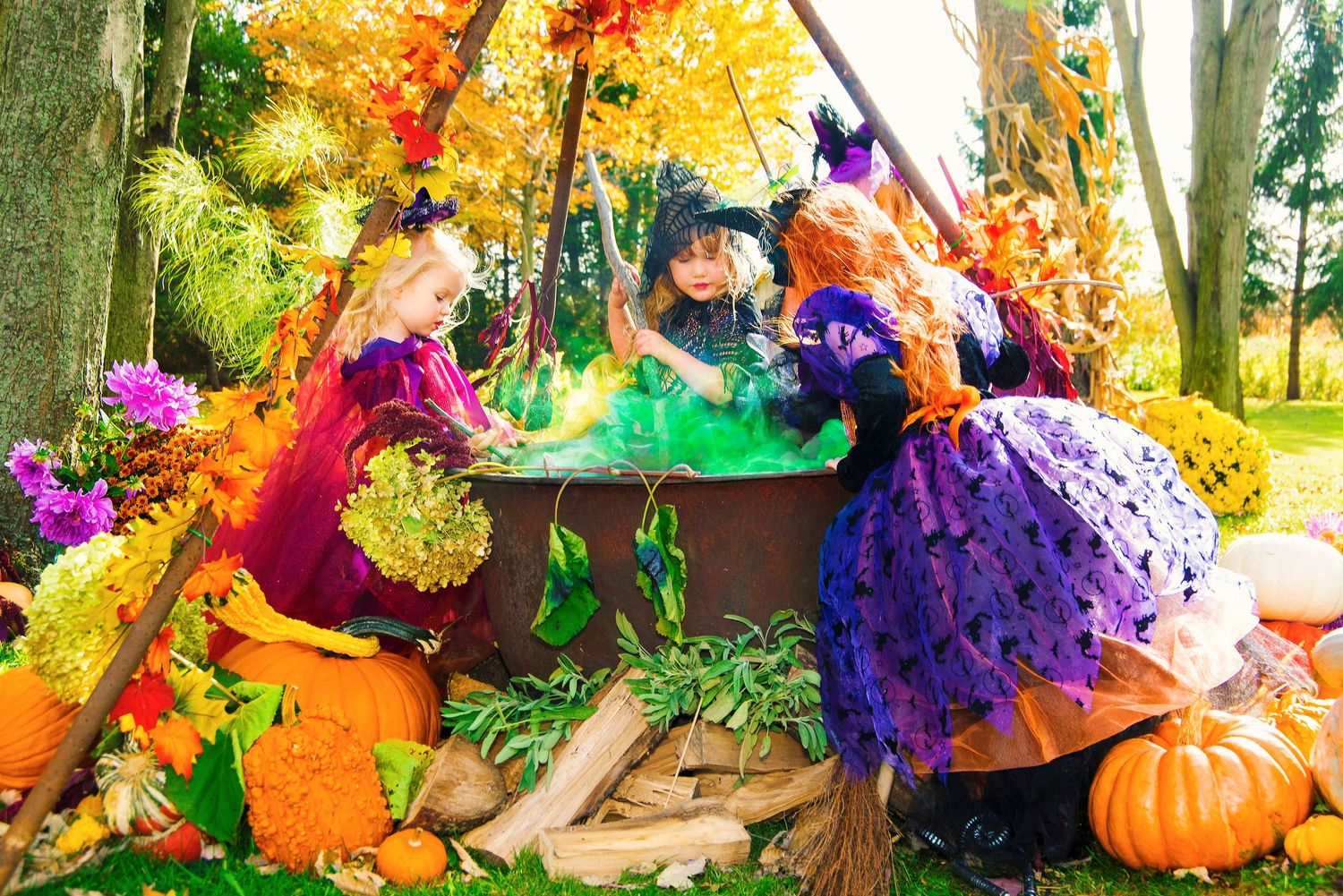 Three girls in witch costumes stirring a smoking cauldron surrounded by autumn Halloween decorations.