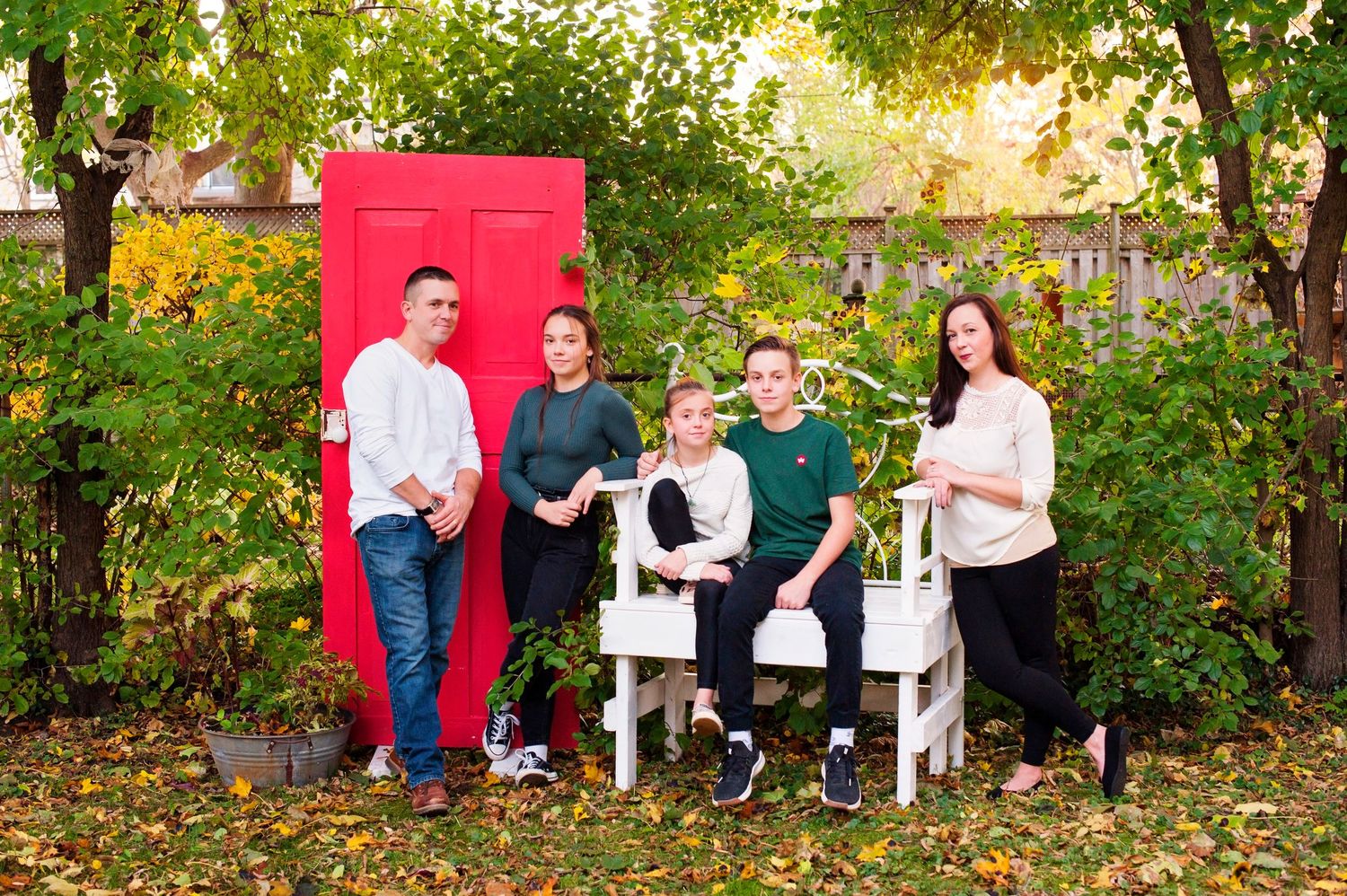 Family of five posing outdoors near a red door surrounded by autumn foliage.