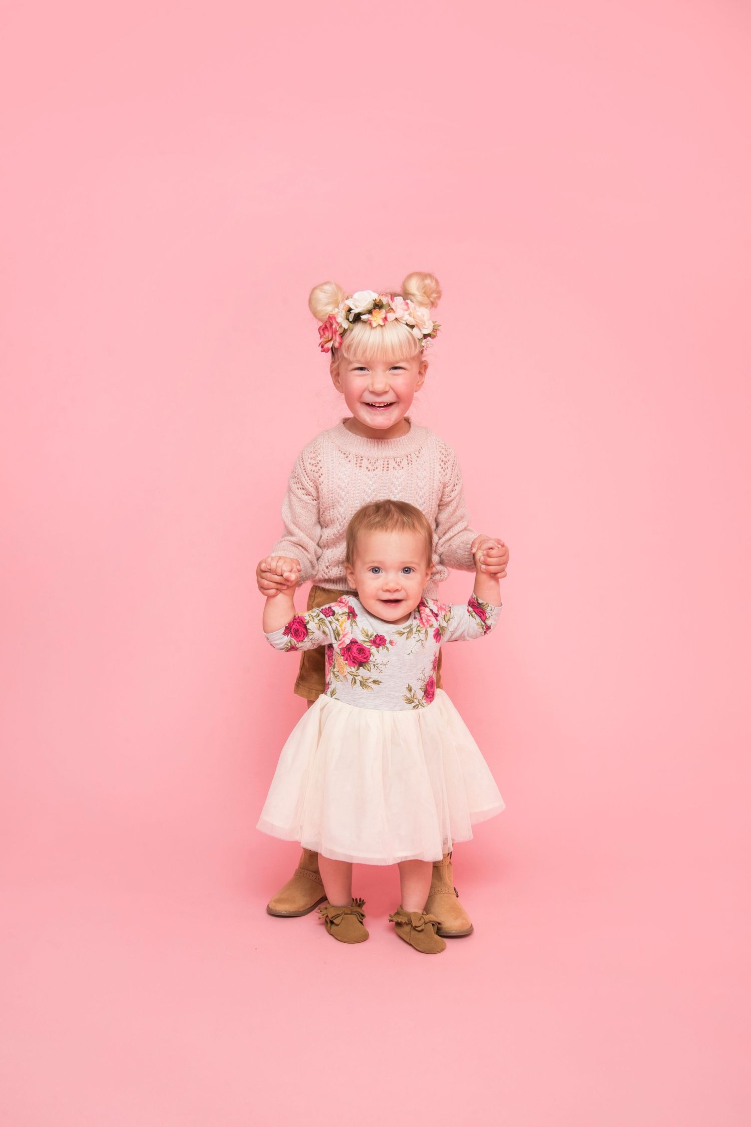 Two young sisters in pink outfits smiling together against a soft pink studio background.