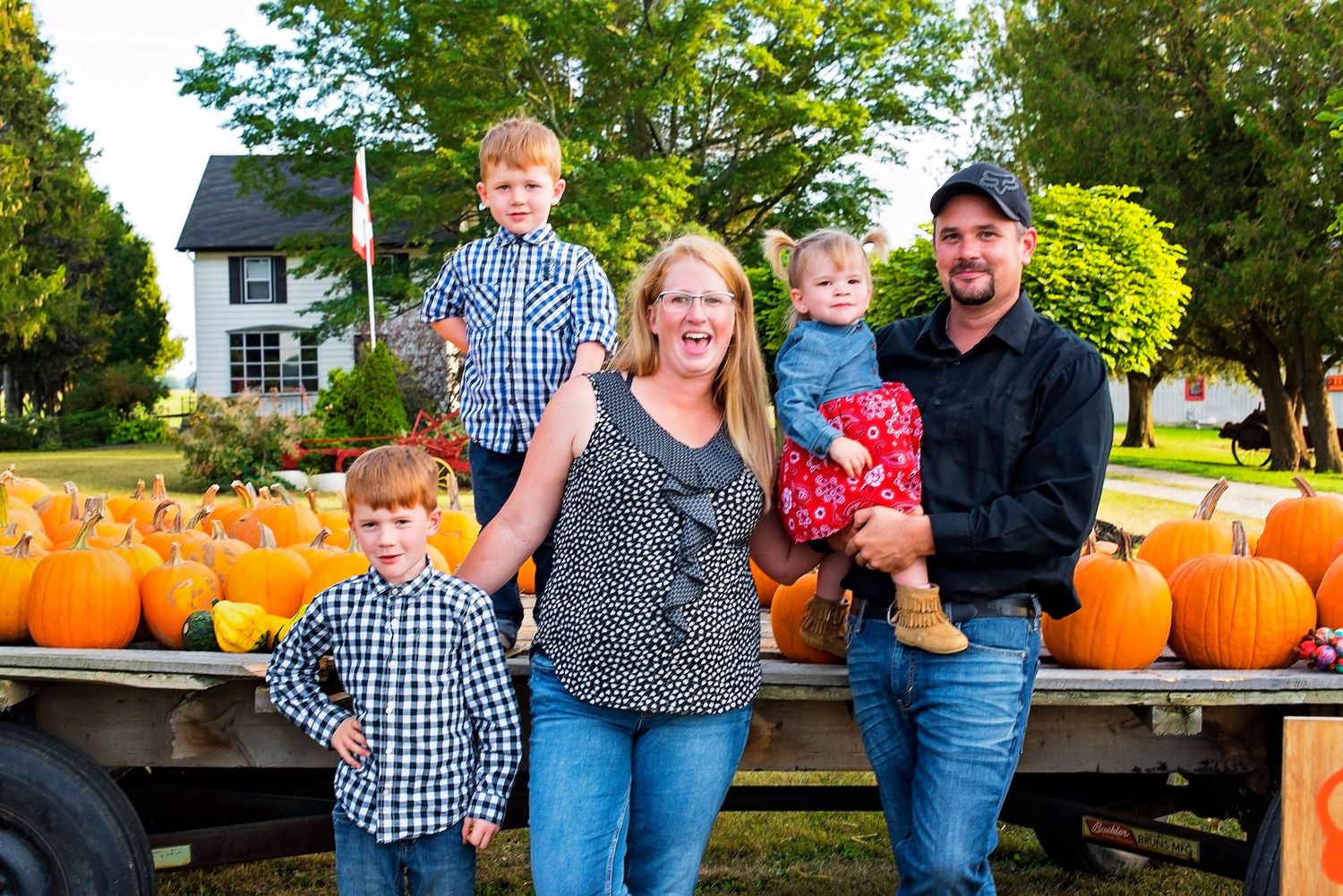 Smiling family of five posing in front of a trailer loaded with orange pumpkins at harvest.