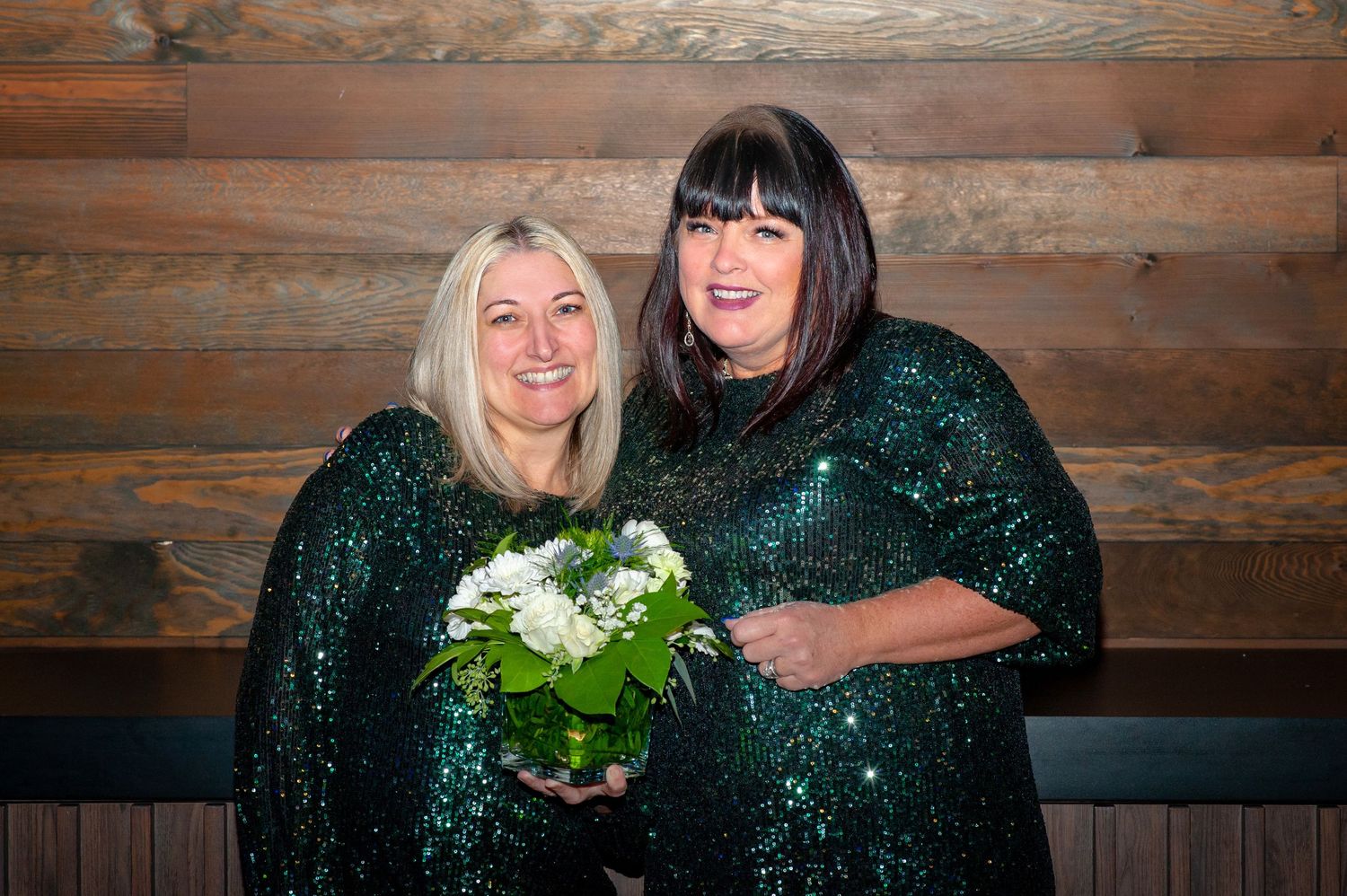 Two women in matching green sequin dresses pose together holding white flowers.