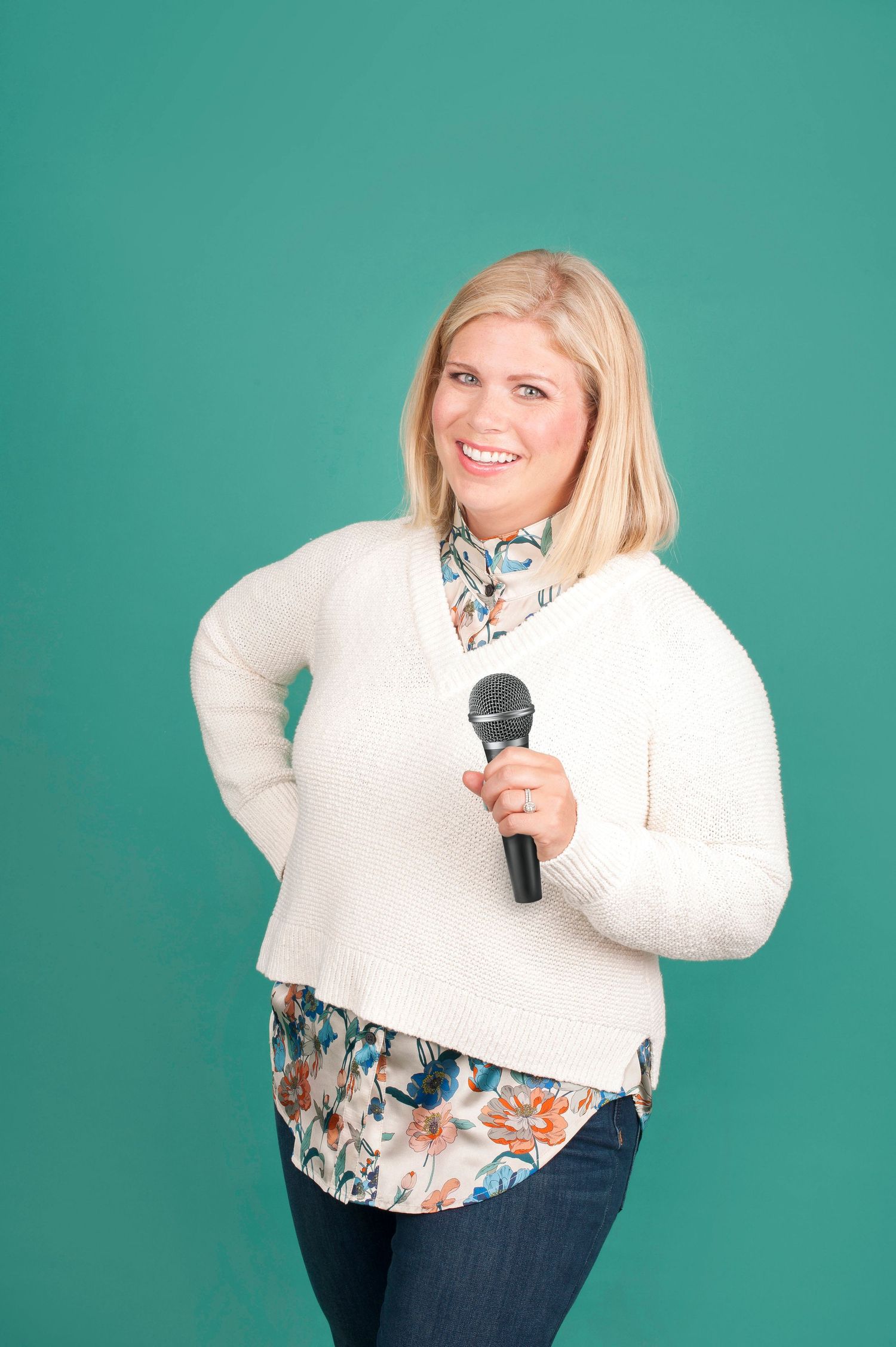 Smiling blonde woman holding a microphone wearing a white cardigan and floral shirt against a teal background.