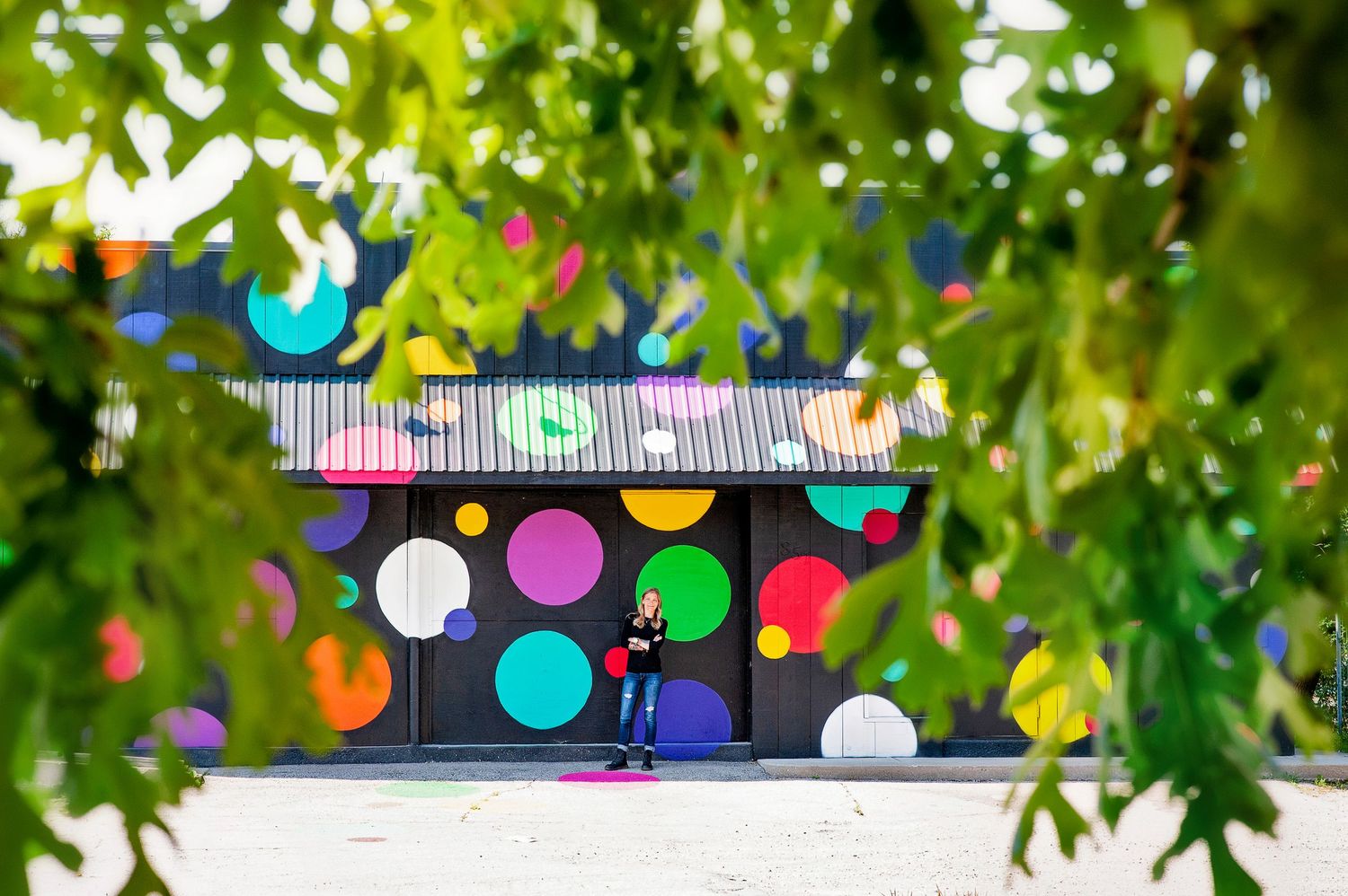 Person standing before a building decorated with colorful polka dot mural, framed by green leaves.