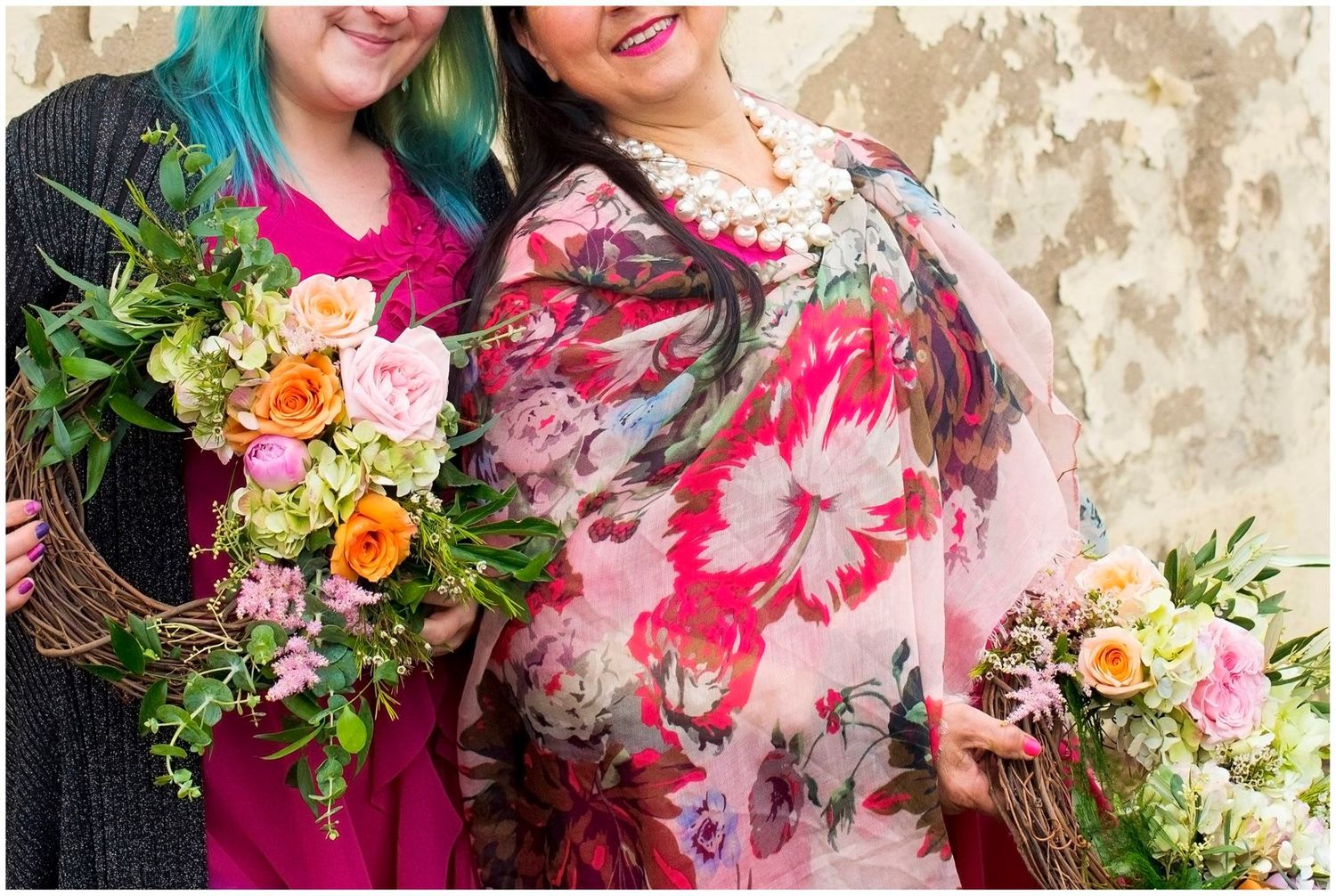 Two women smiling and holding colorful floral bouquets against a rustic textured wall background.