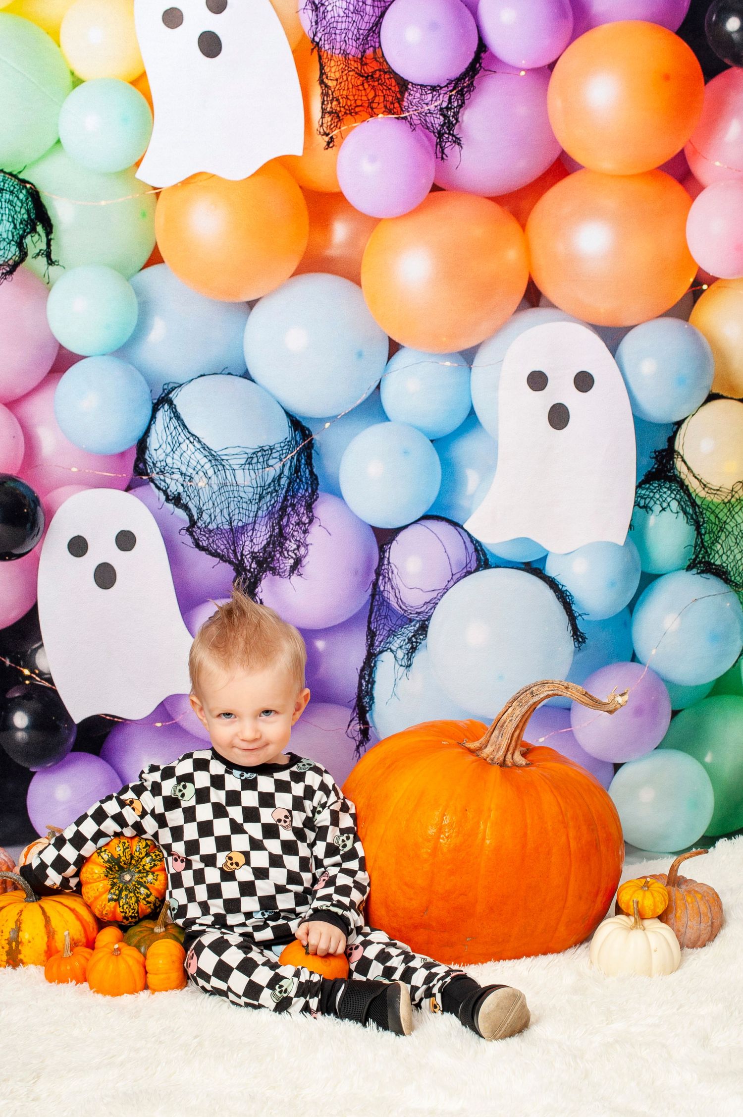 Toddler boy in checkered Halloween outfit sits with pumpkins against colorful balloon wall with ghost decorations.