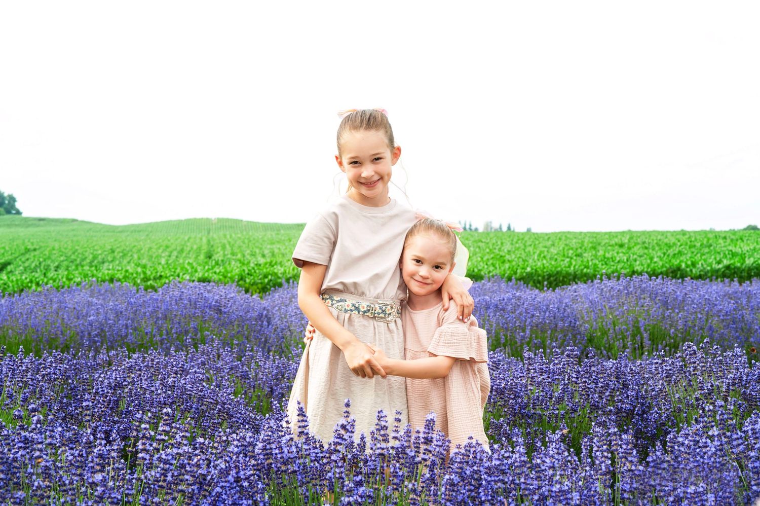 Two smiling sisters in beige dresses hug each other while standing in a lush purple lavender field.
