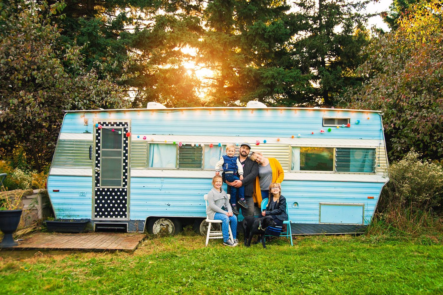 Family gathered outside retro blue travel trailer during warm golden hour sunset.