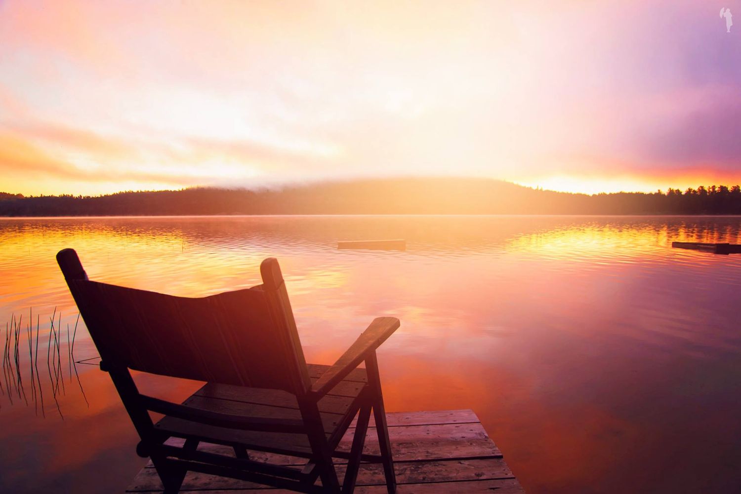 Wooden chair on a dock overlooking a calm lake during a stunning pink and orange sunset.