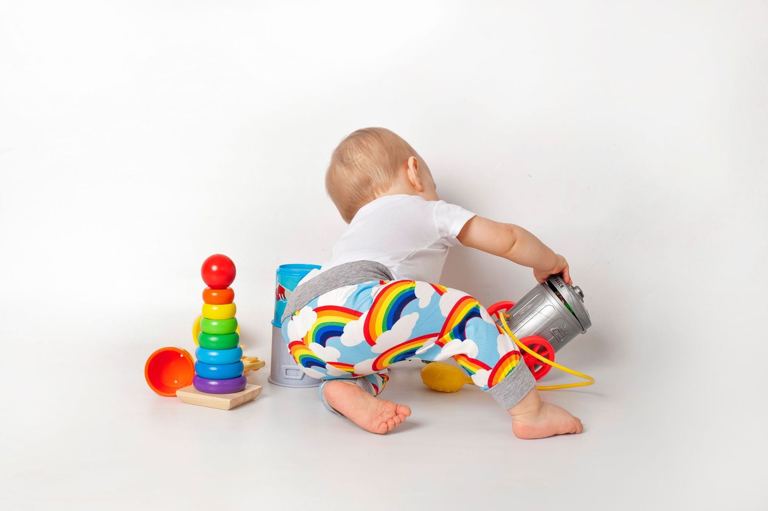 Baby in rainbow pants playing with colorful stacking toys on a clean white background.