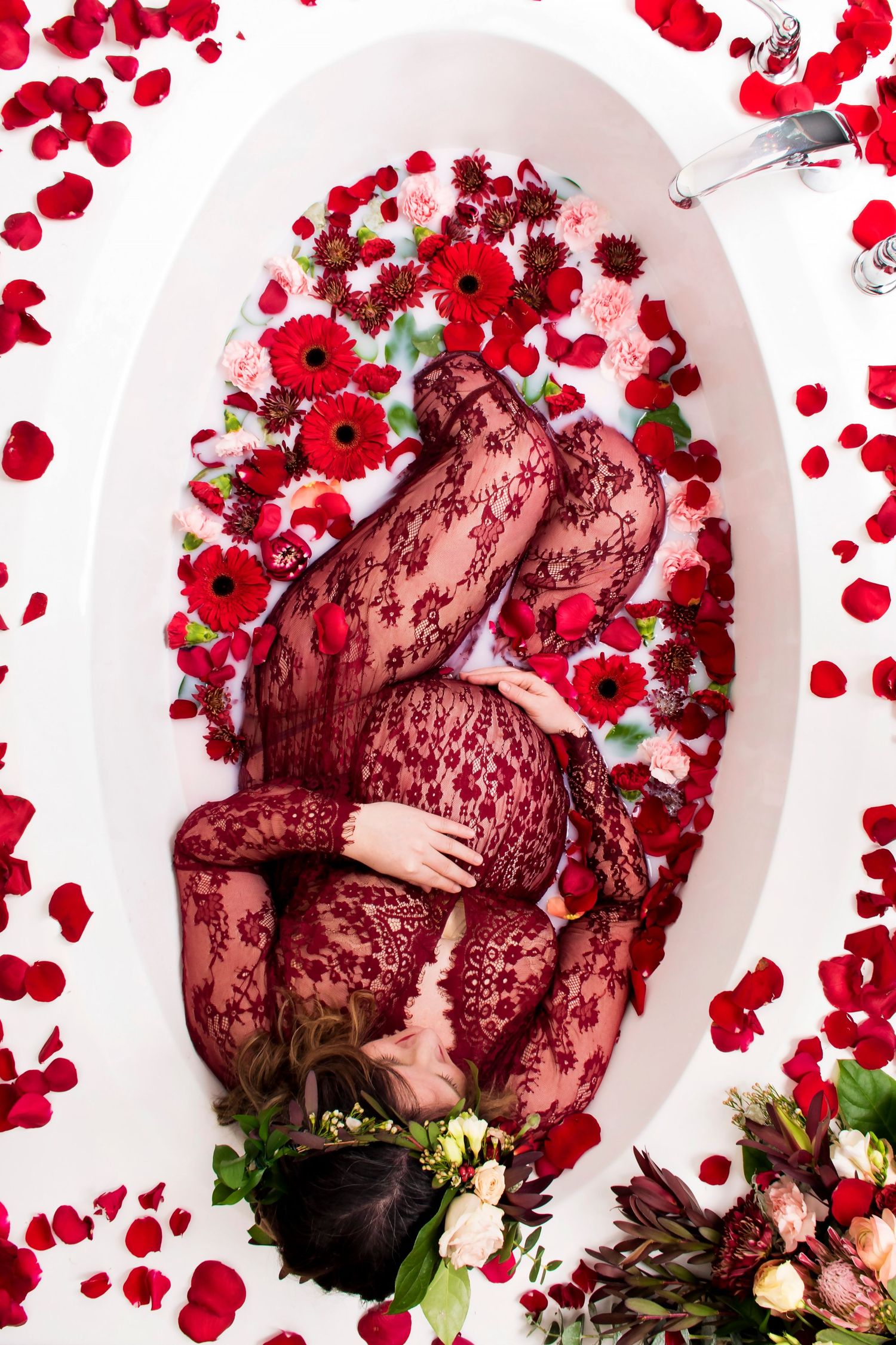 Pregnant woman in red lace bodysuit lying in floral bath surrounded by red poppies and rose petals.