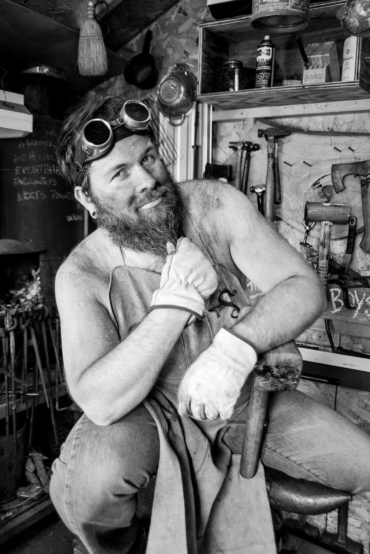 Bearded shirtless man with goggles and work gloves resting in a rustic workshop, black and white portrait.